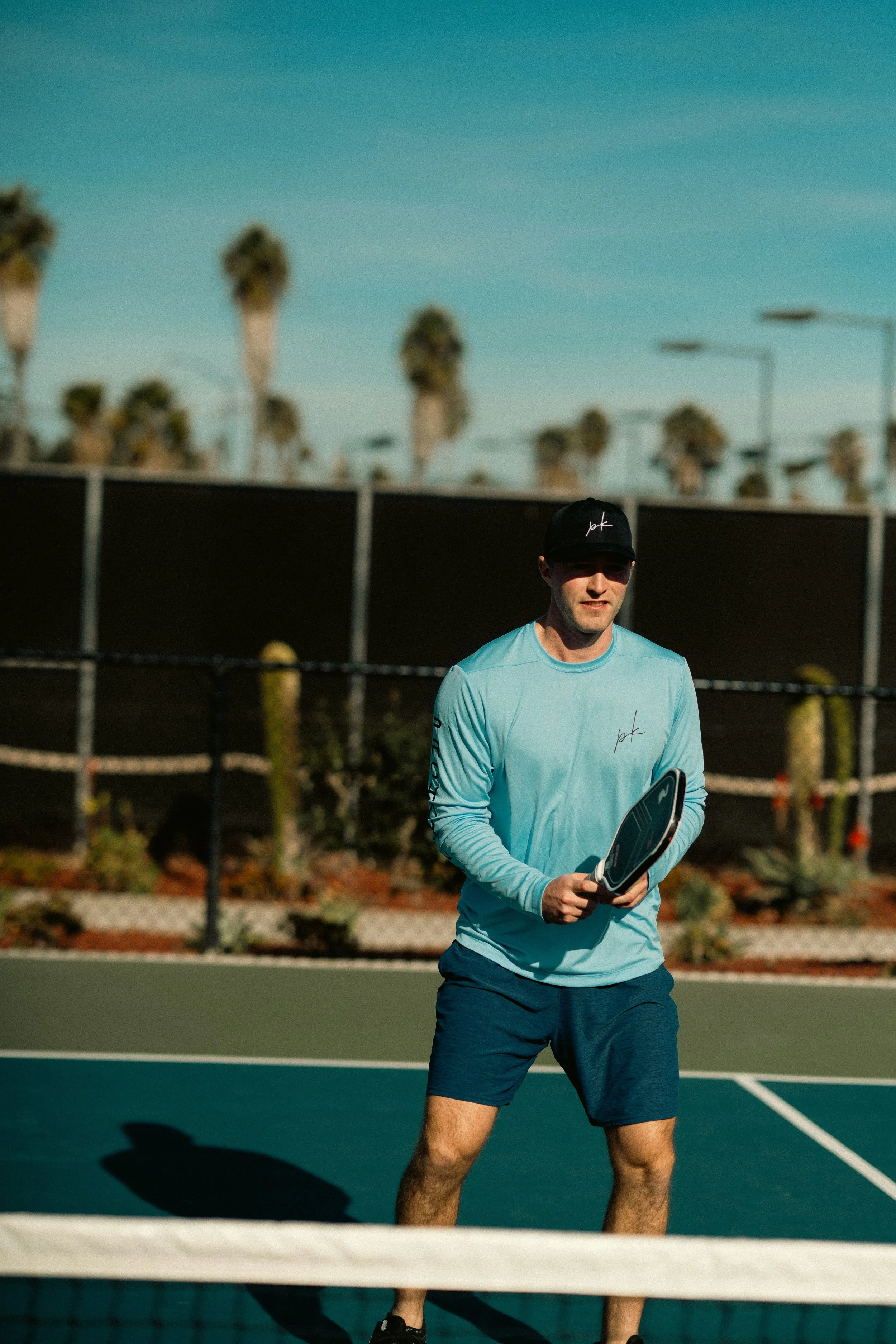 A man playing pickleball on an outdoor court during daytime, wearing a black cap, light blue long-sleeve shirt, and navy shorts, holding a paddle.