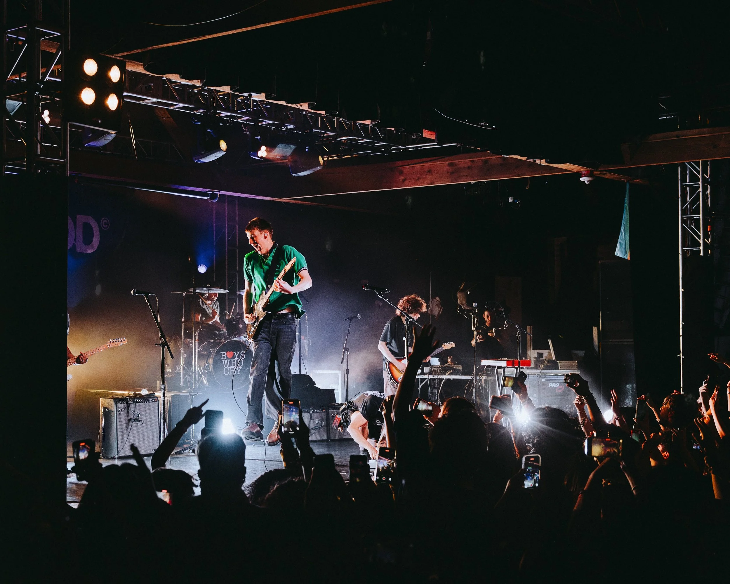 A live band performs on stage with a crowd taking photos and videos, bright stage lights illuminate the performers, and some band members are playing guitars while others are on drums and keyboard.
