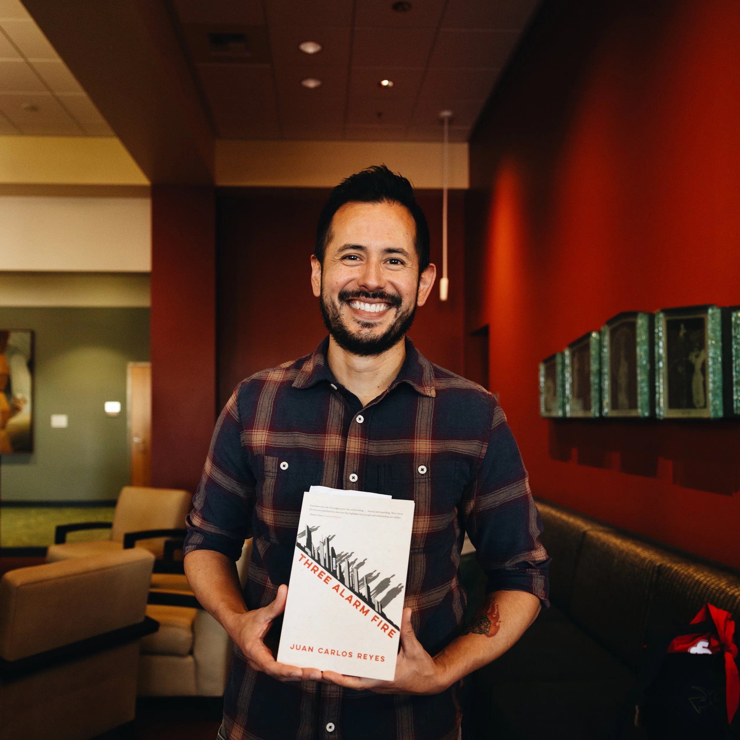 A smiling man with dark hair and a beard holding a book titled 'Three Alarm Fire' by Juan Carlos Reyes, standing in a warmly lit room with red walls and artwork on the wall.