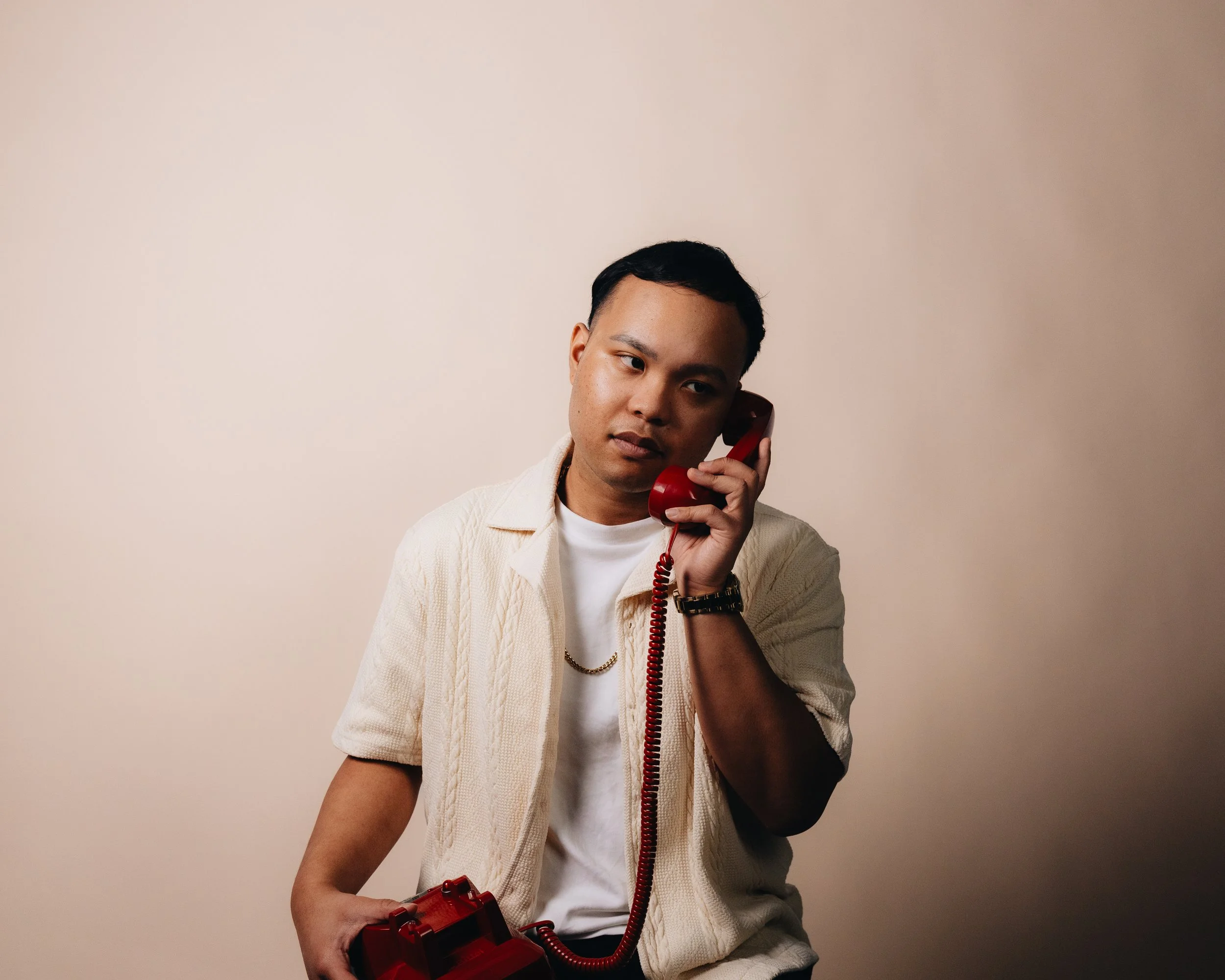 A young man seated against a plain beige wall, holding a red rotary phone to his ear with a serious expression.