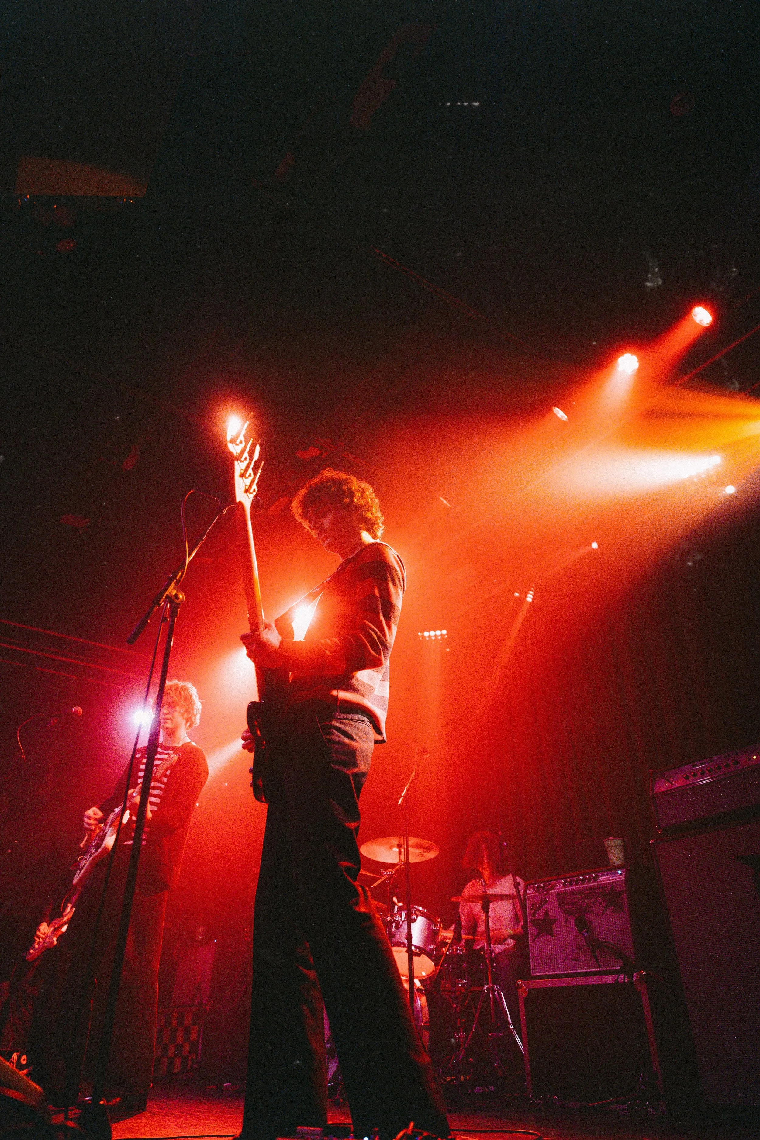 Band performing on stage with orange and red stage lighting, featuring a guitarist in the foreground, a bassist to the left, and a drummer in the background.