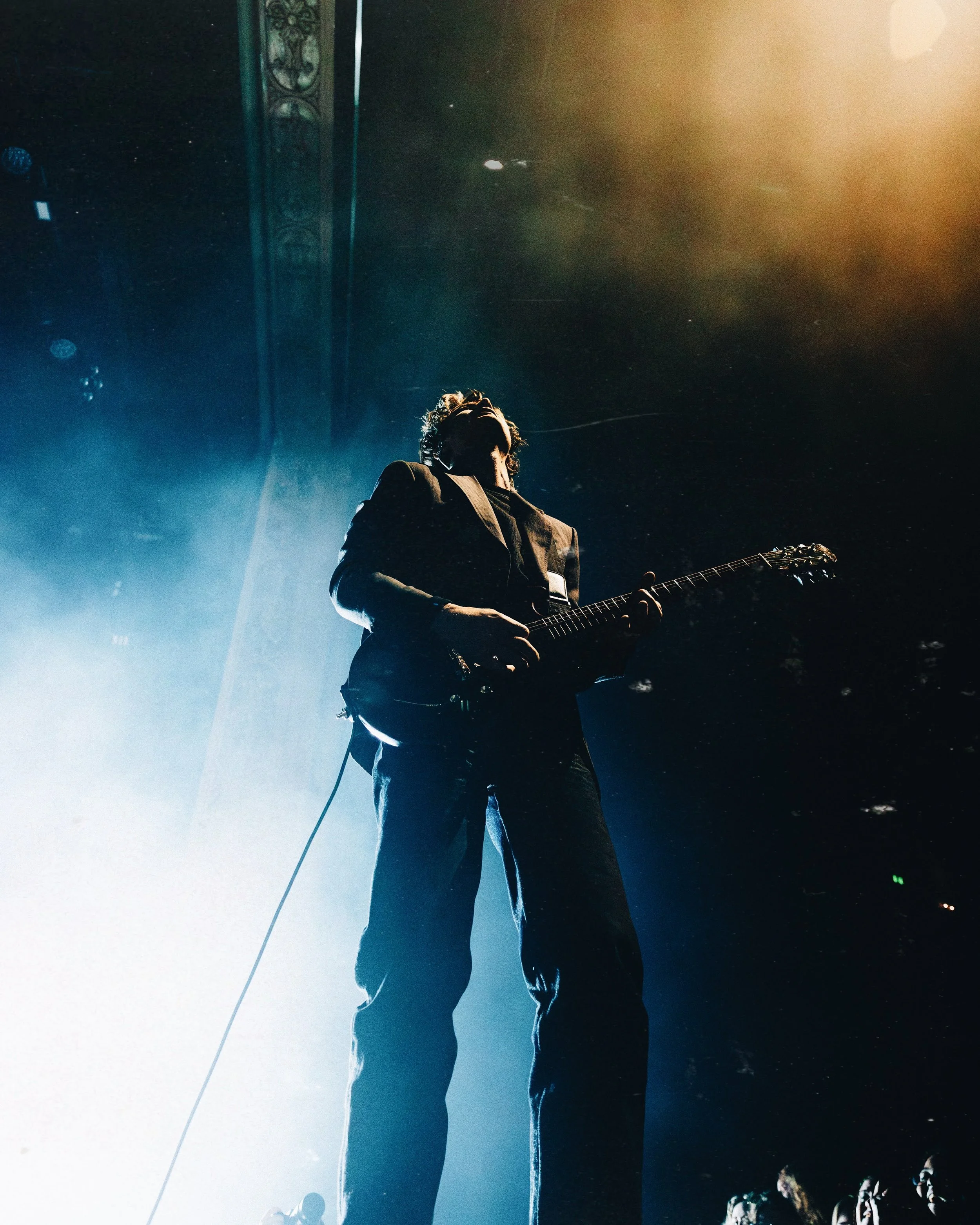 A musician playing an electric guitar on stage with dramatic lighting and an audience in the background.