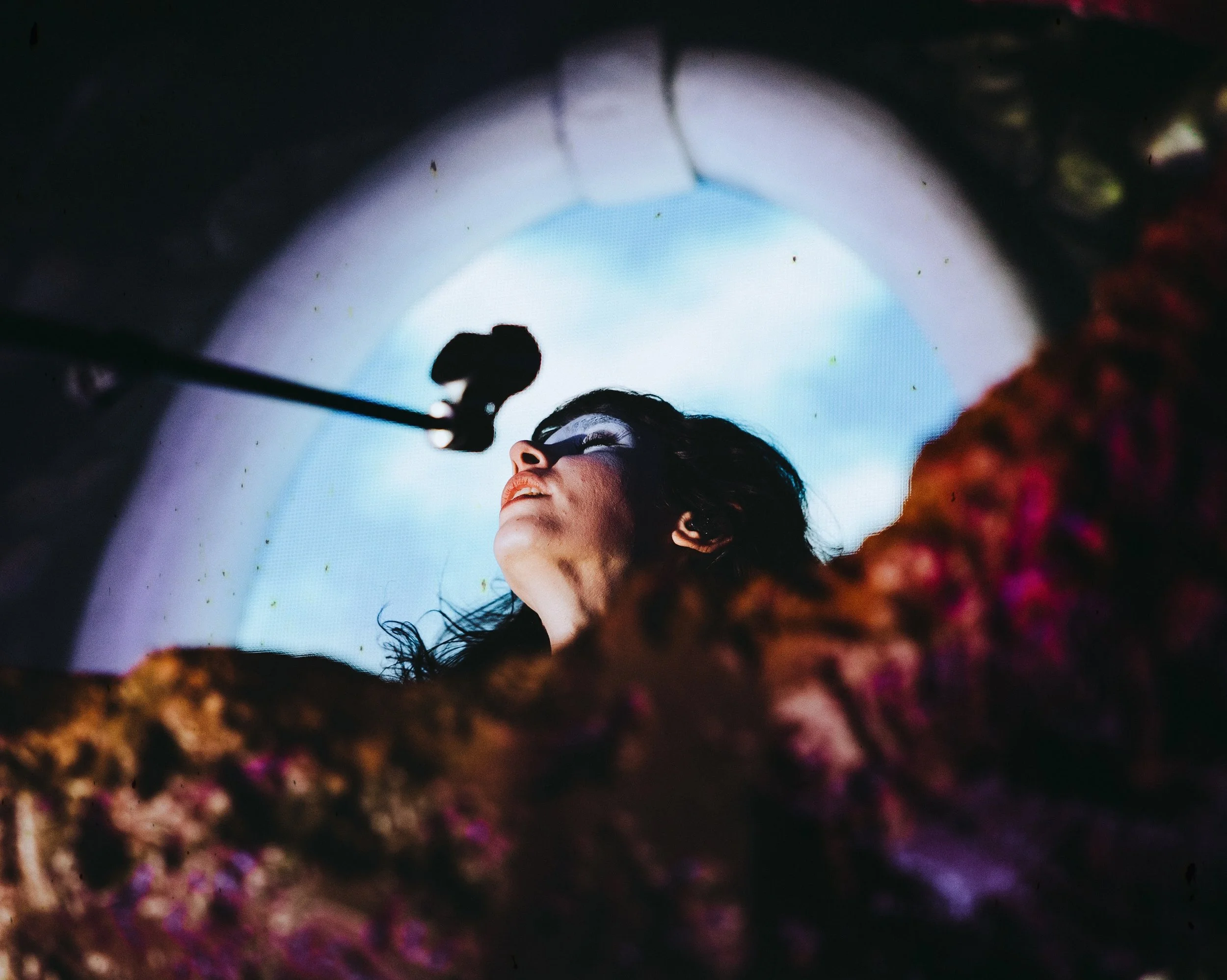 A woman with dark hair and makeup looking upward, seen from a low angle inside a circular opening with a cloudy sky visible in the background.