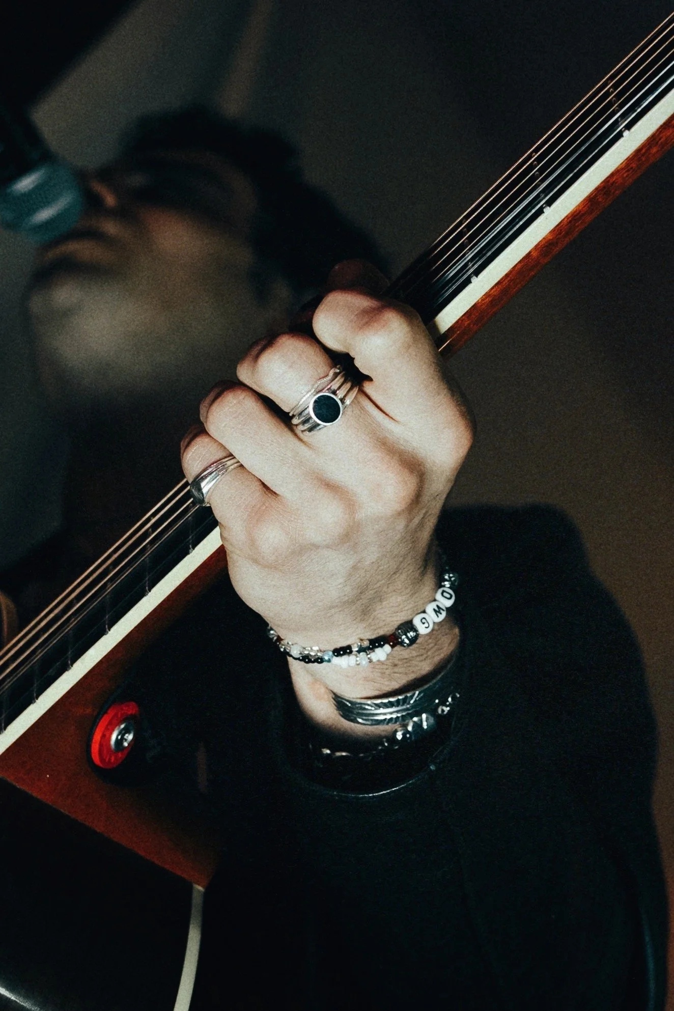 Close-up of a person's hand with rings and bracelets, holding a guitar neck, reflected in a mirror, with a bottle and water in the background.
