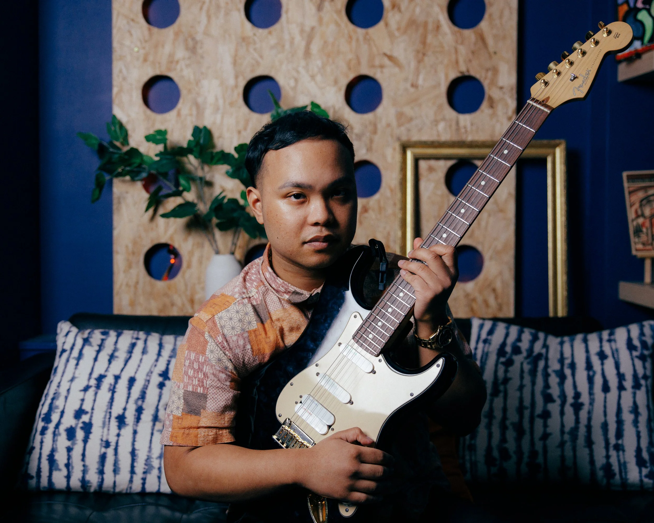 A young man holding a black and white electric guitar in a room with blue walls, a wooden decorative panel, and a couch with striped pillows.
