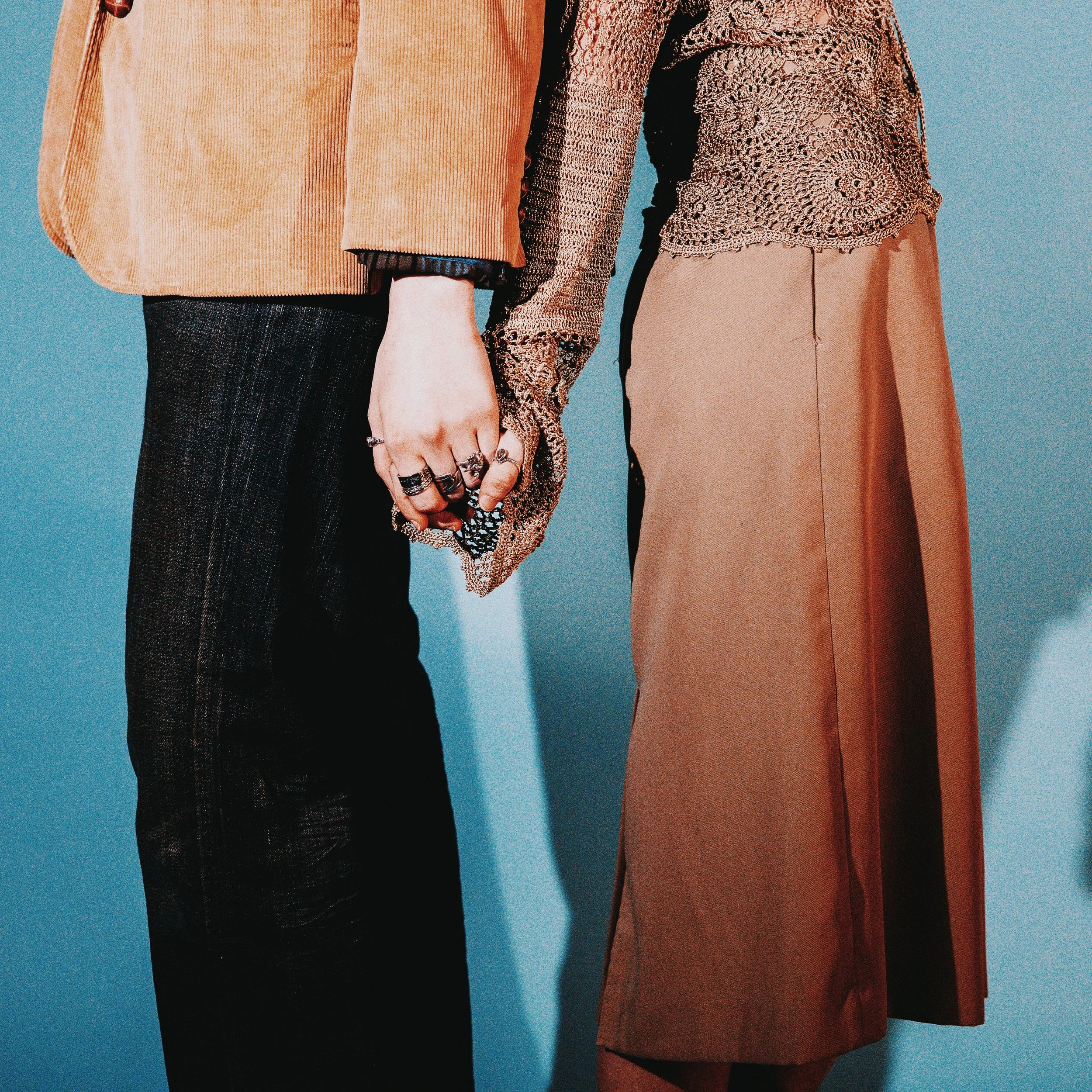 Two people holding hands, showing their clothing and jewelry. One person is wearing a tan blazer, black pants, and rings, while the other is wearing a brown skirt and a textured blouse.
