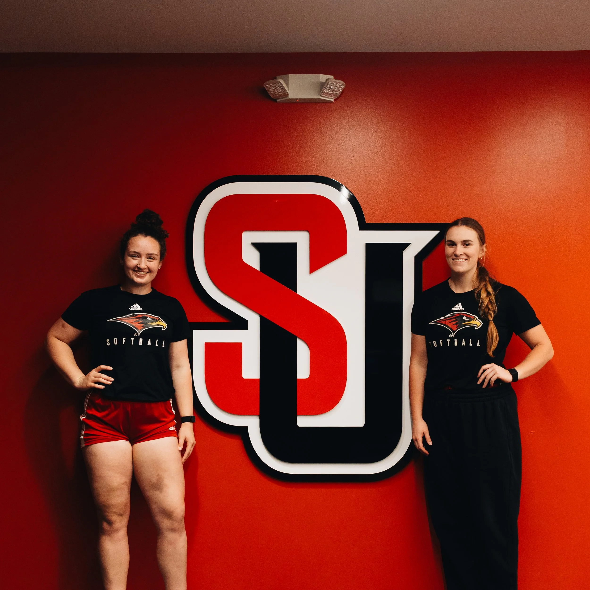 Two women in softball uniforms standing in front of a red wall with a large black, red, and white logo of a stylized letter 'S' and 'U',