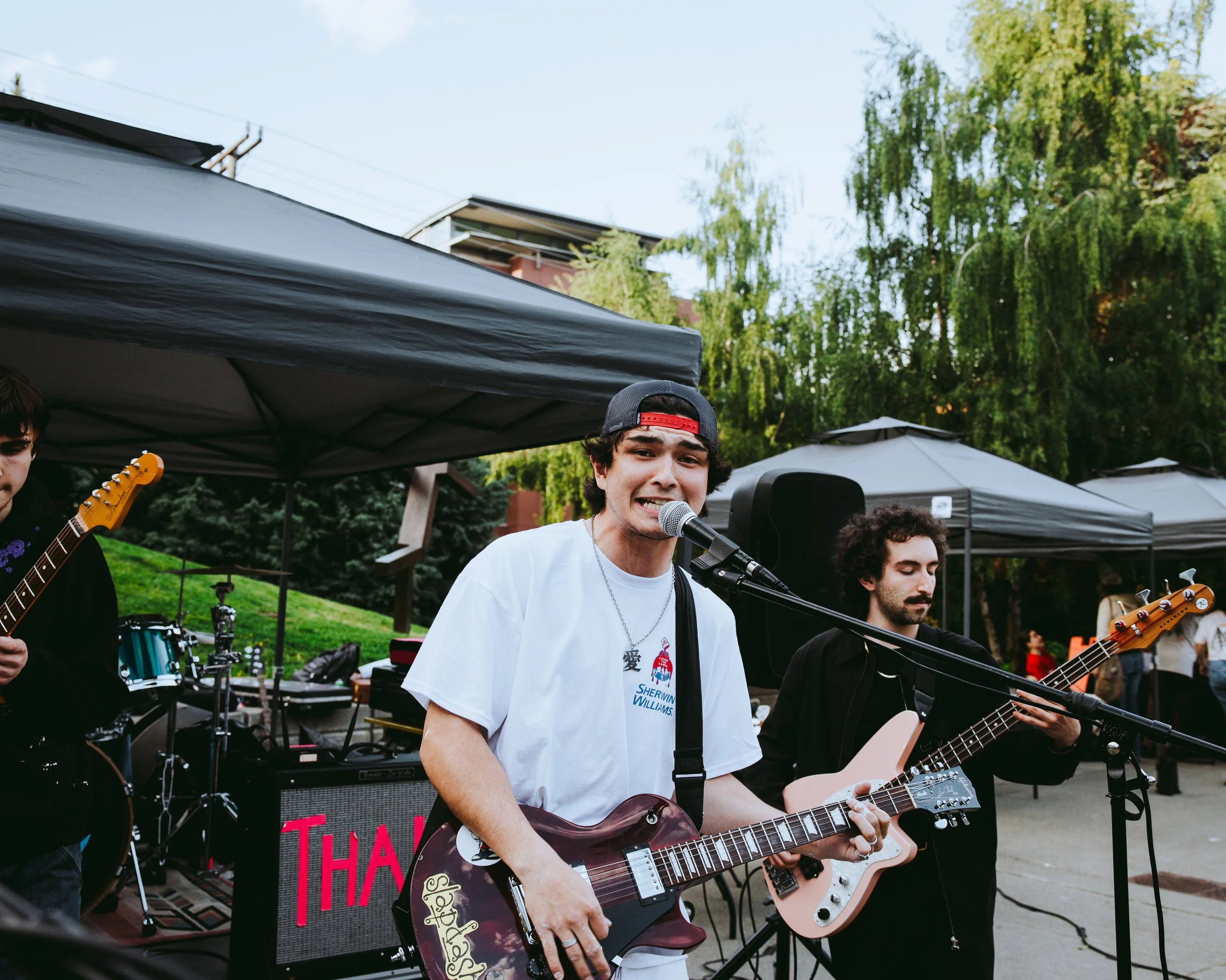 A young man wearing a white t-shirt and a backwards black cap is playing an electric guitar and singing into a microphone at an outdoor music event. There are two other band members with guitars beside him and tents in the background.