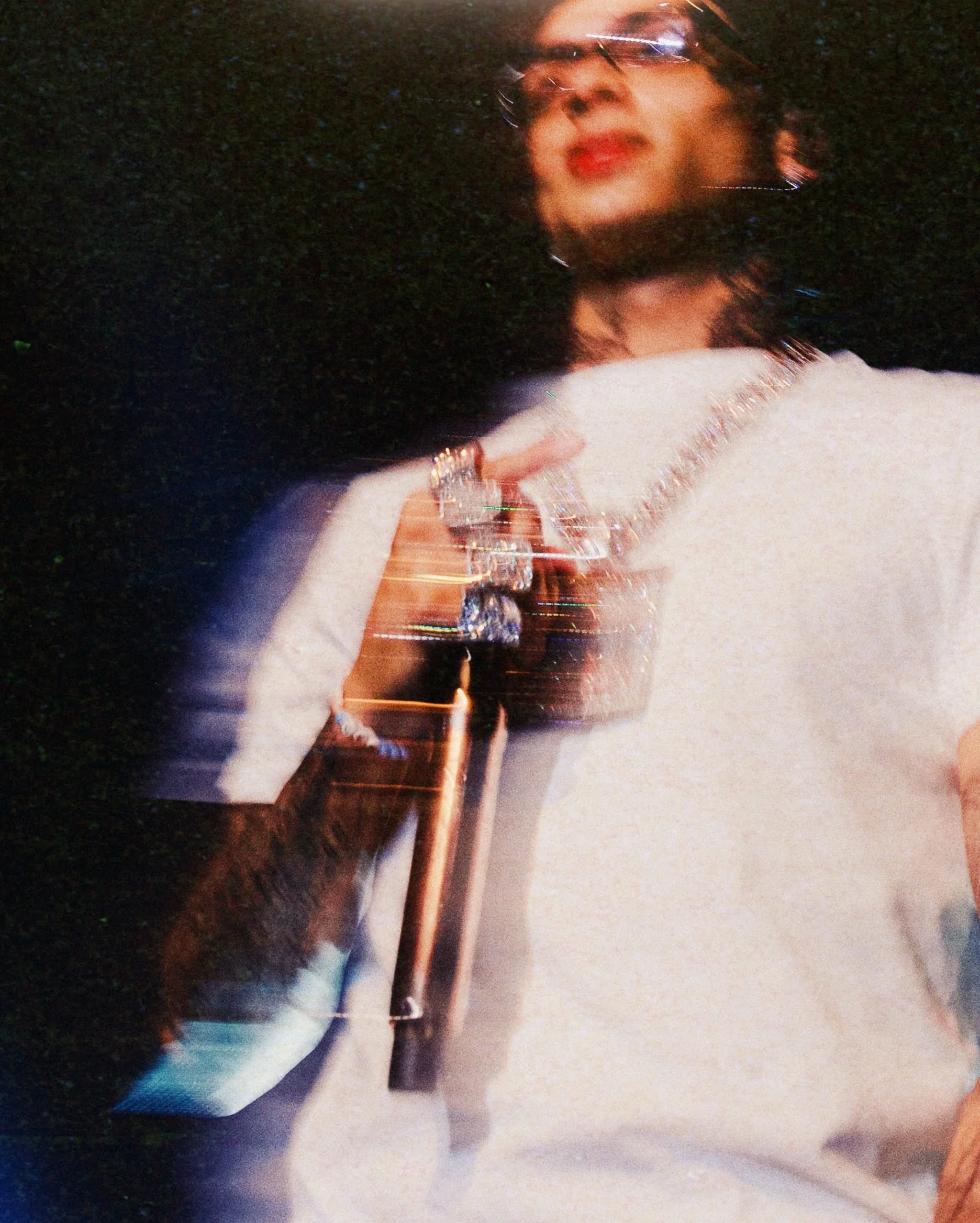A woman with dark hair, wearing glasses, a white sweater, and jewelry, standing outdoors at night with a dark background, captured with motion blur and light streaks.