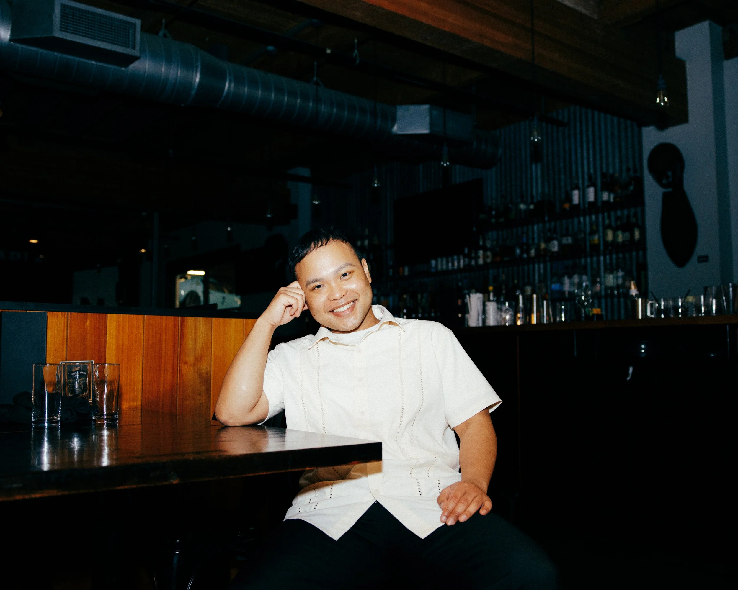 A smiling man sitting at a bar in a dimly lit restaurant or bar, resting his head on his right hand while looking at the camera.