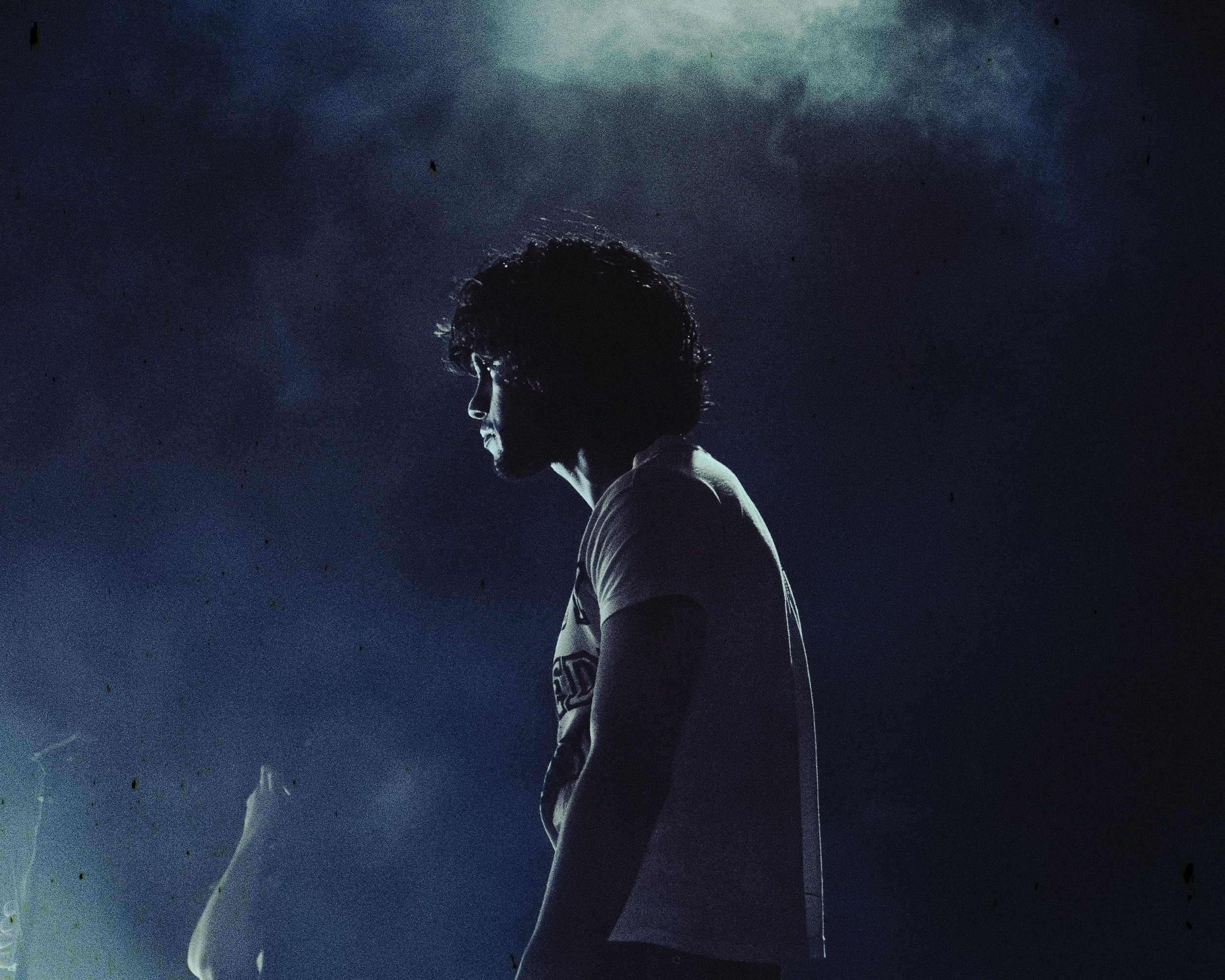 A person with curly hair standing in profile against a dark, cloudy background with subtle lighting.