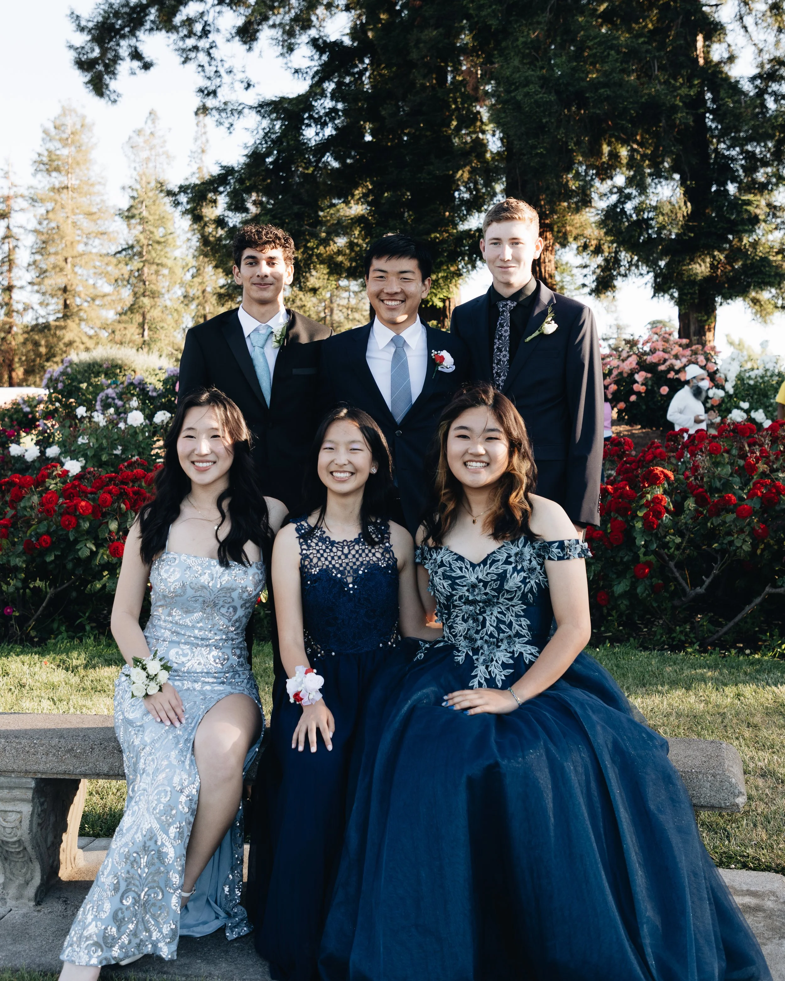 Group of six young people dressed in formal attire, sitting and standing in a garden with colorful flowers and trees in the background.