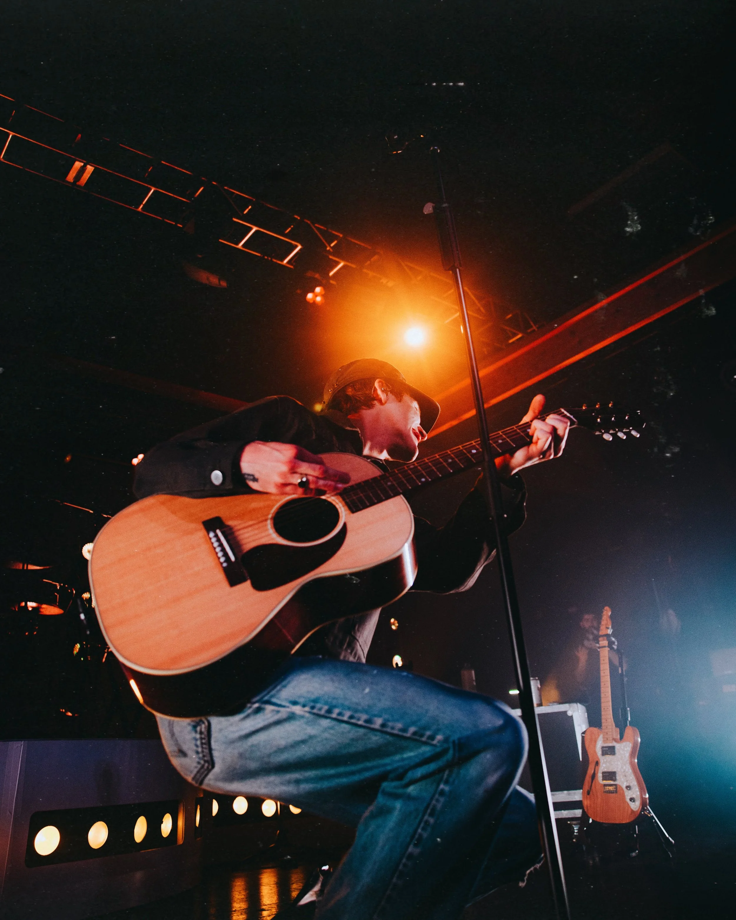 A musician playing an acoustic guitar on stage with stage lights and another guitar in the background.