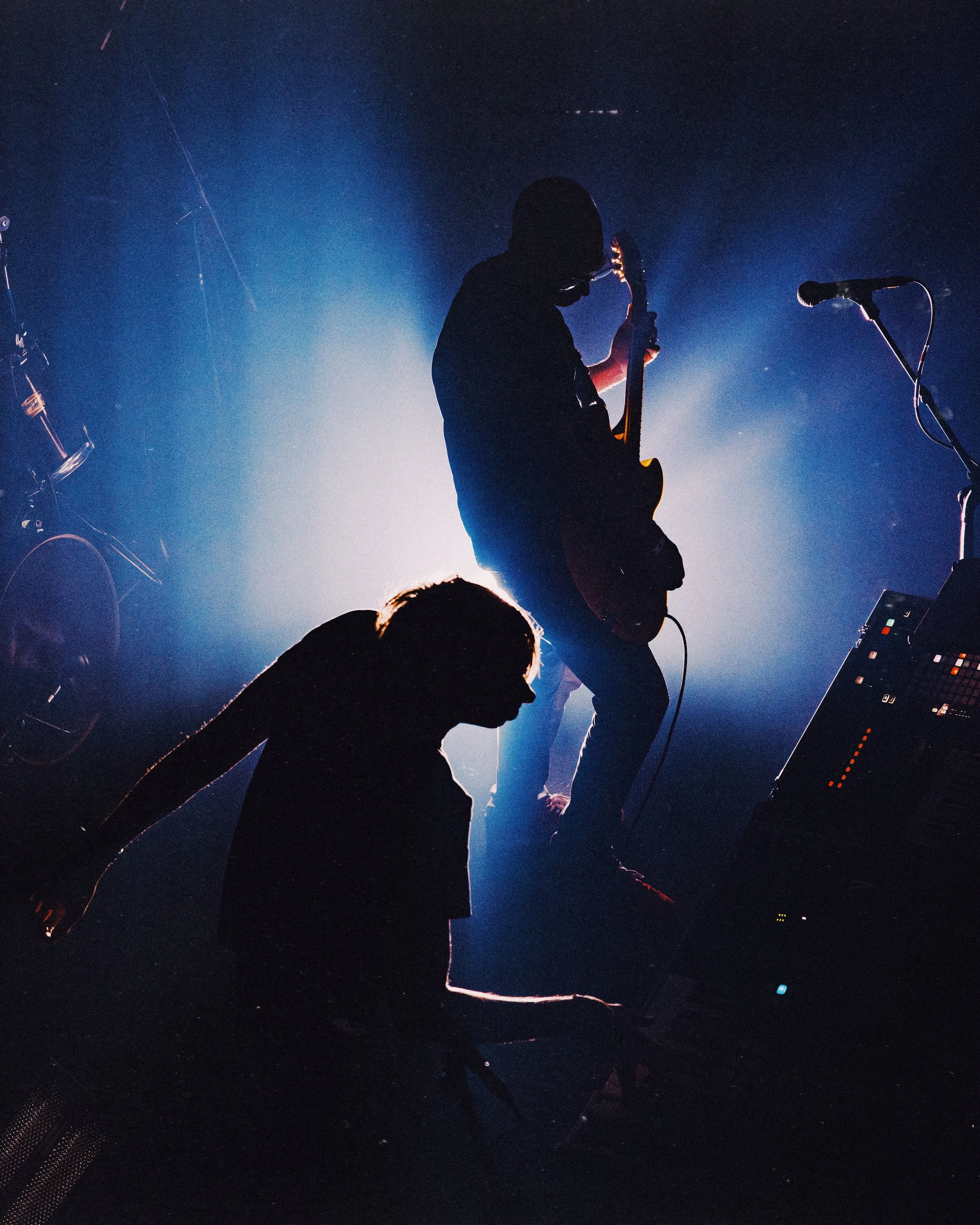 Silhouettes of two musicians performing on stage, one playing an electric guitar and the other at a keyboard, with bright blue lighting and stage equipment around.