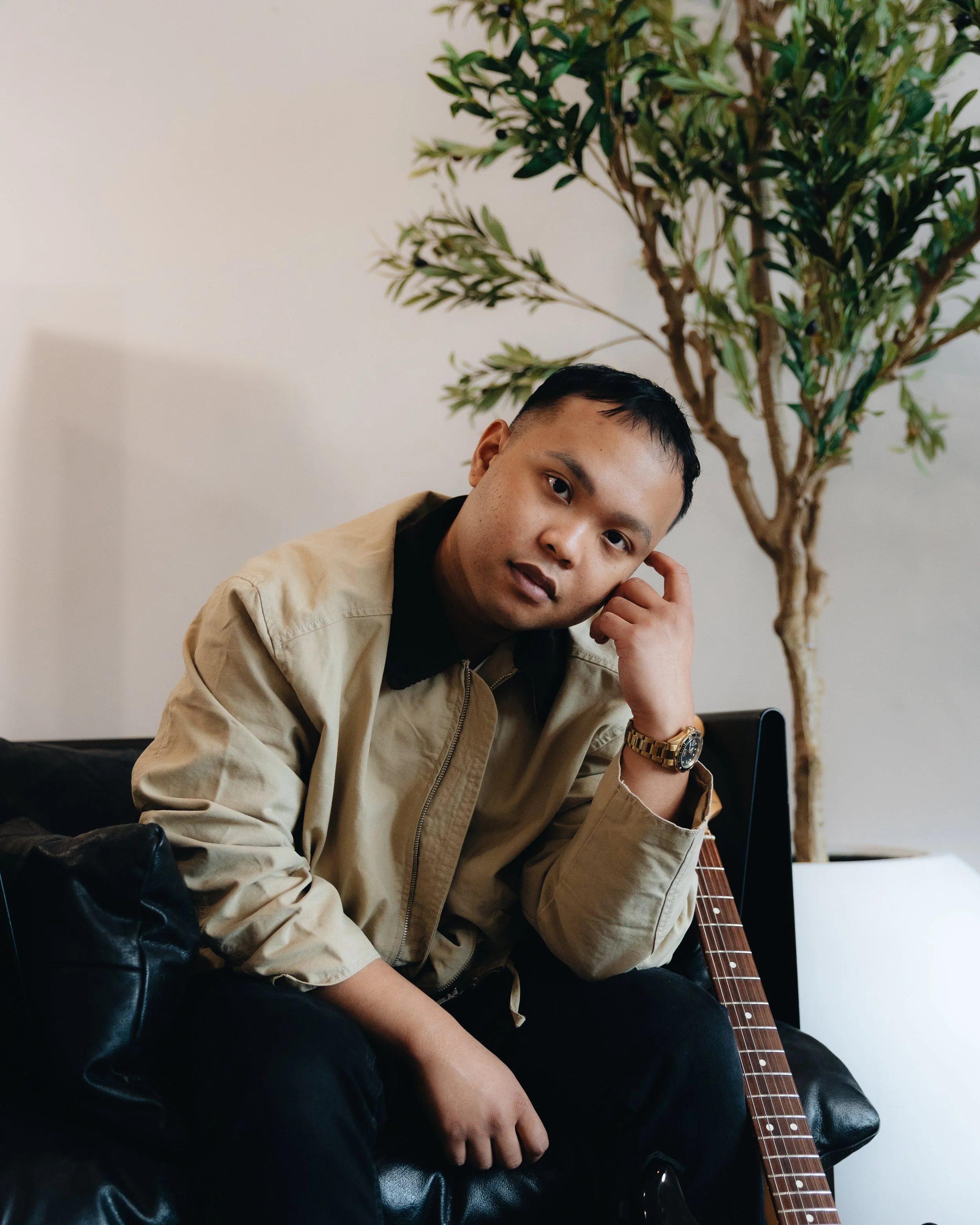 A young man sitting on a black leather couch with an acoustic guitar beside him, touching his face, wearing a beige jacket and a gold watch, with a tall green plant in the background.