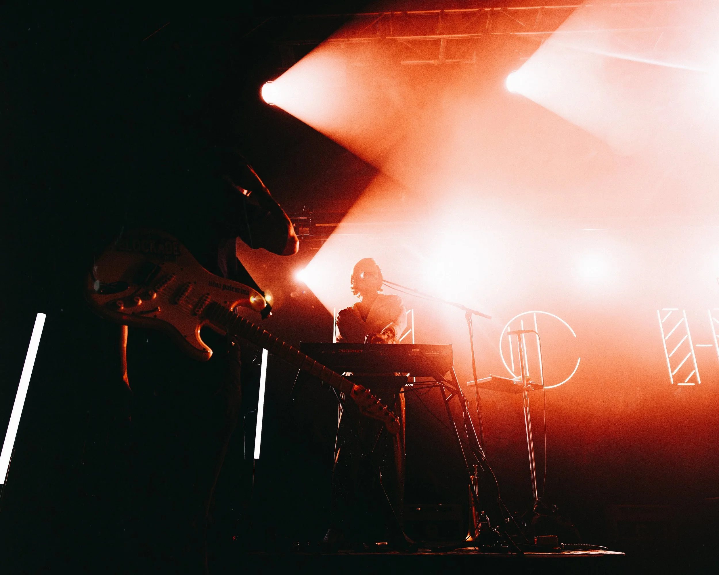 Musicians performing on stage with orange-red lighting, one playing guitar and the other on keyboard, with neon outlines of letters in the background.