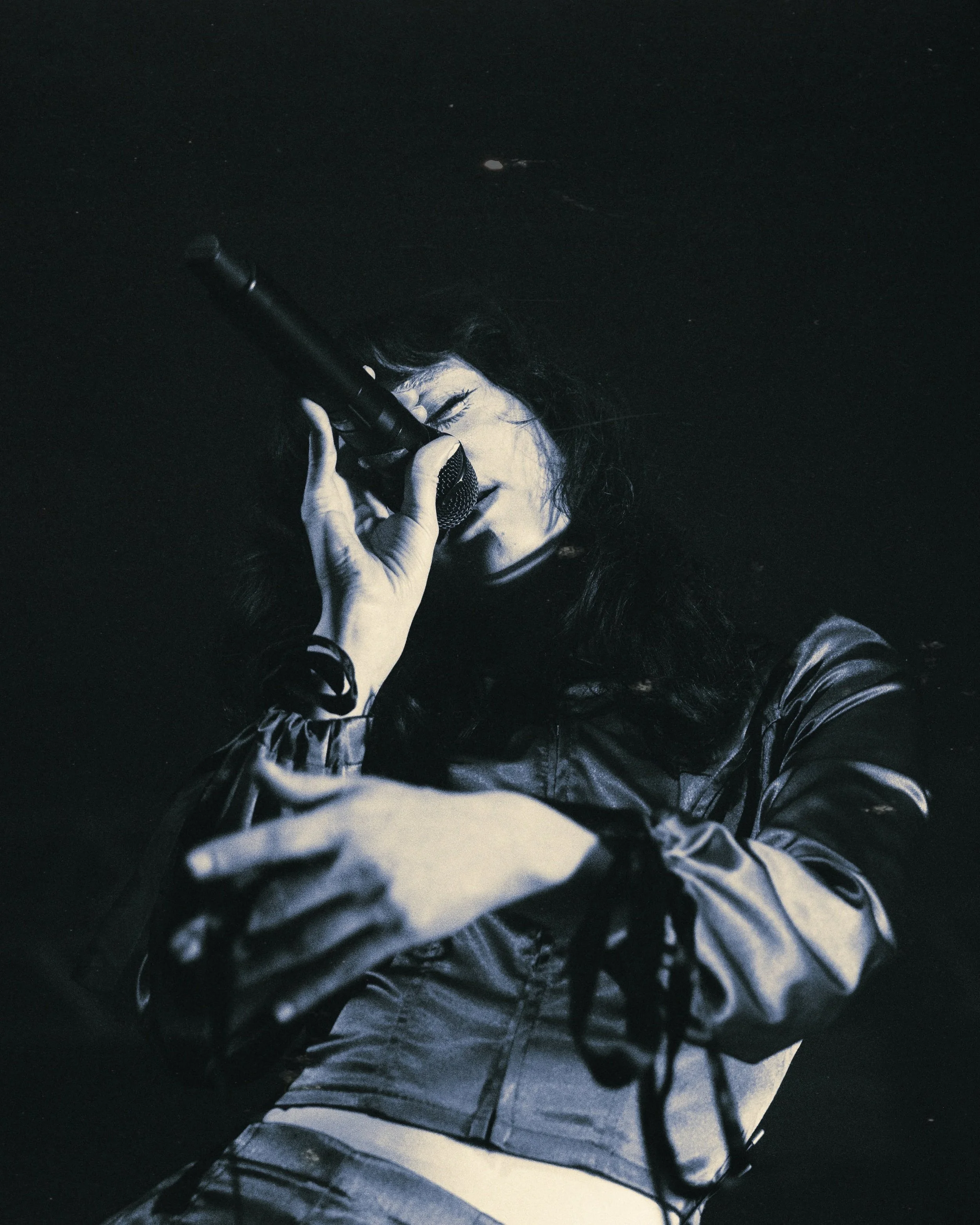 A woman passionately singing into a microphone on stage, with her eyes closed and her hand holding the microphone close to her face. She has long dark hair and is wearing a shiny jacket.