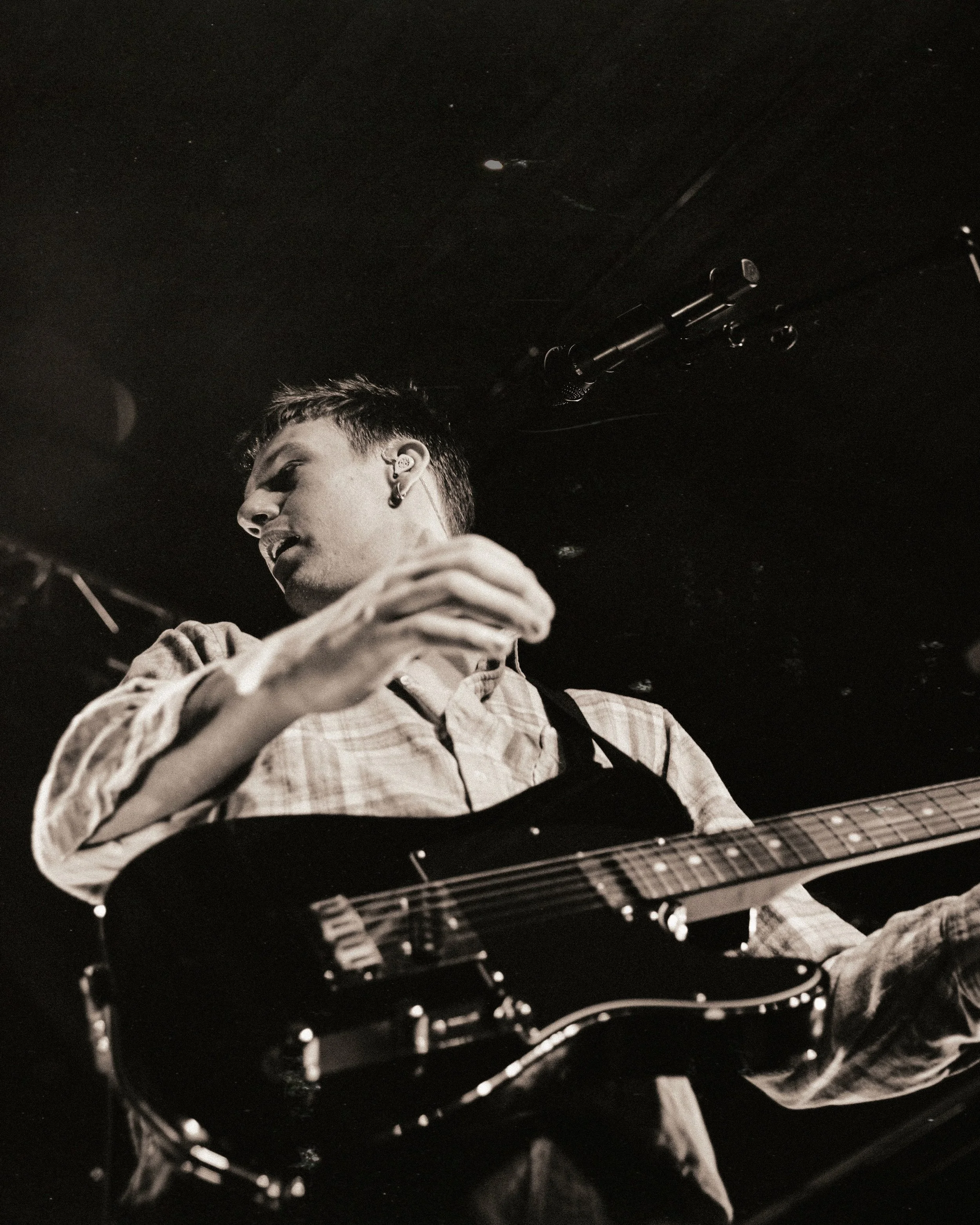 A young man playing an electric guitar on stage, with a microphone above him, in black and white.