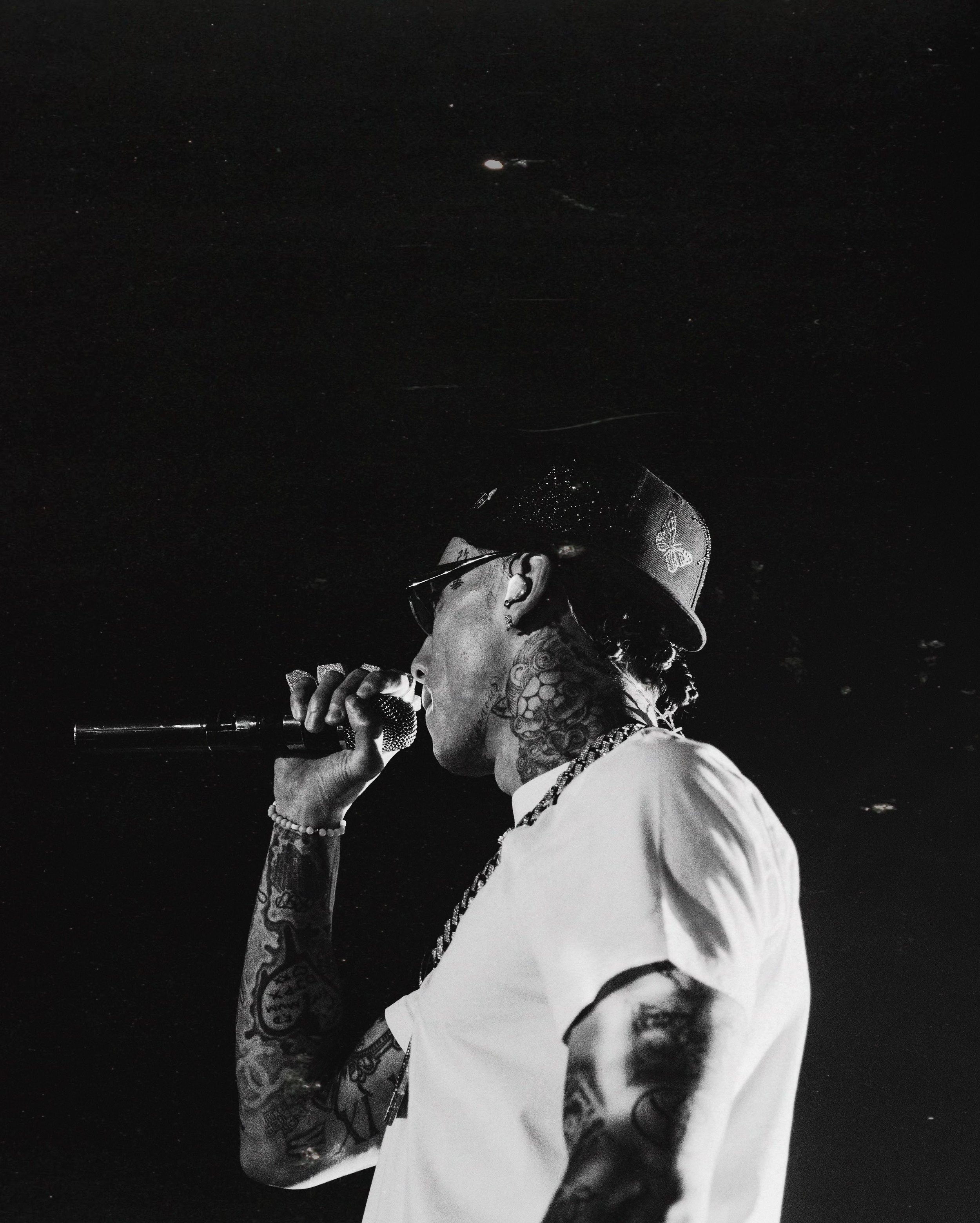A tattooed man singing into a microphone, wearing a baseball cap and sunglasses, in a dark setting.