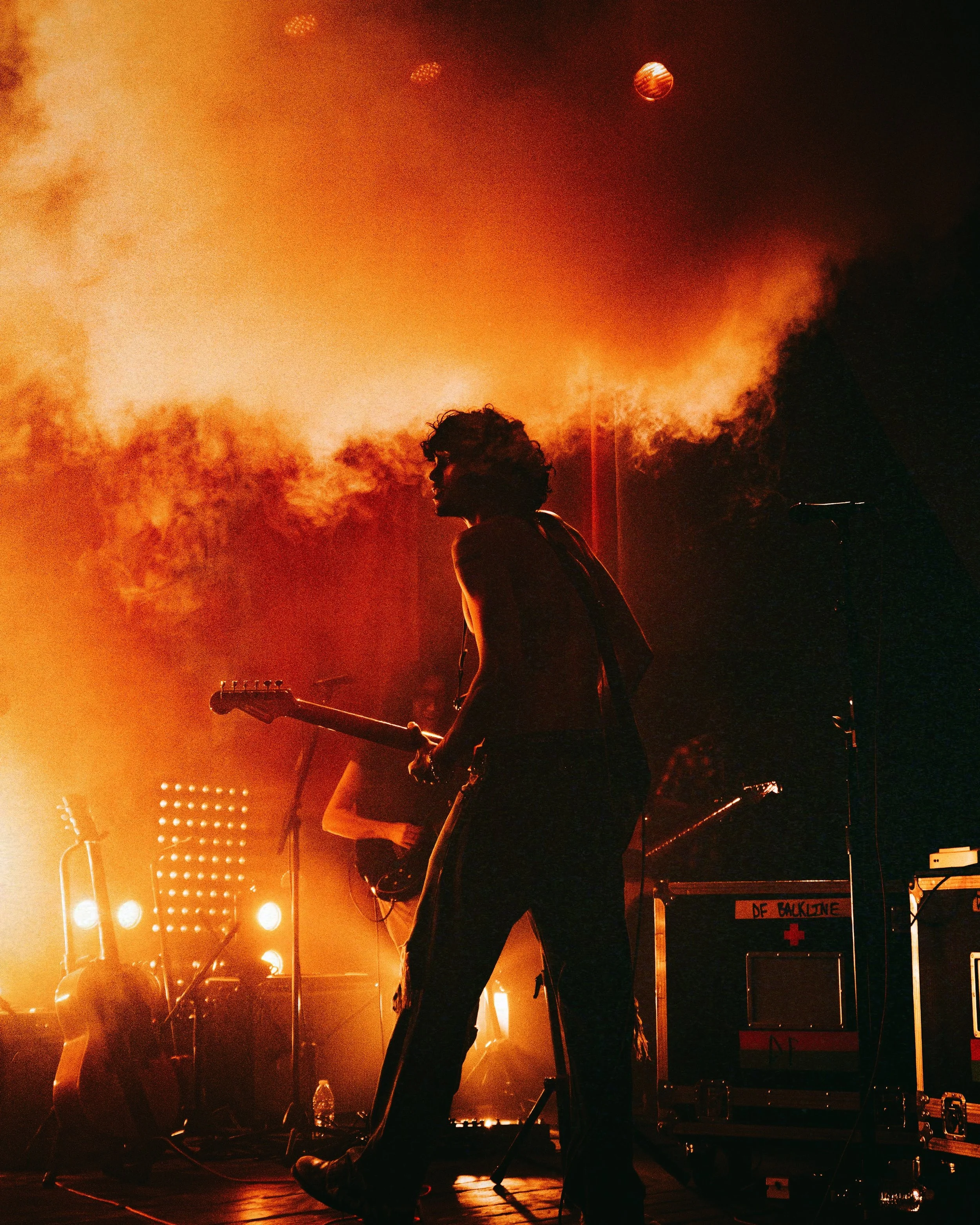 A musician playing guitar on stage with orange lighting, smoke, and several guitars visible in the background.