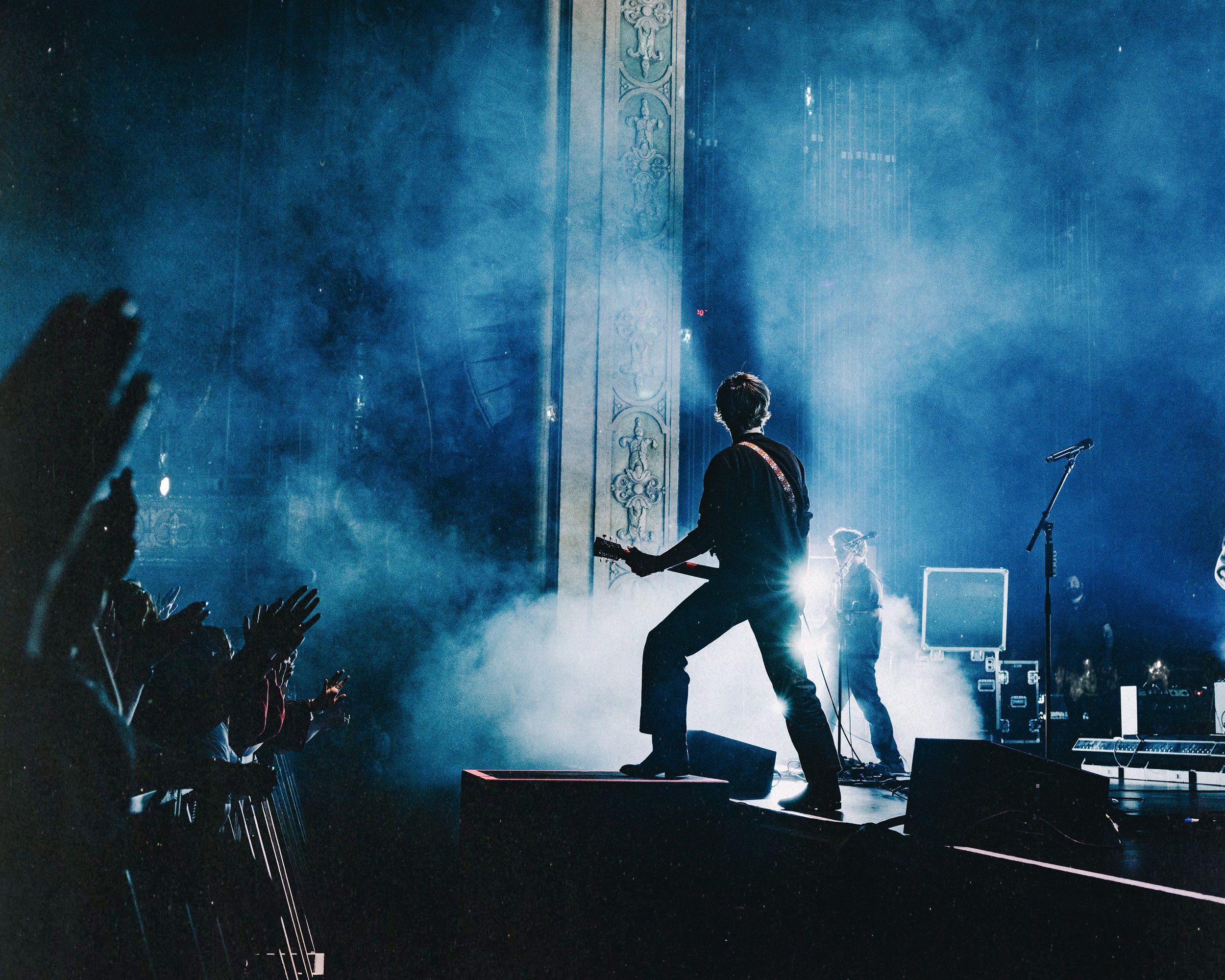 Musician playing guitar on stage during a concert, with audience reaching out and cheering, stage lighting, and smoke effects.