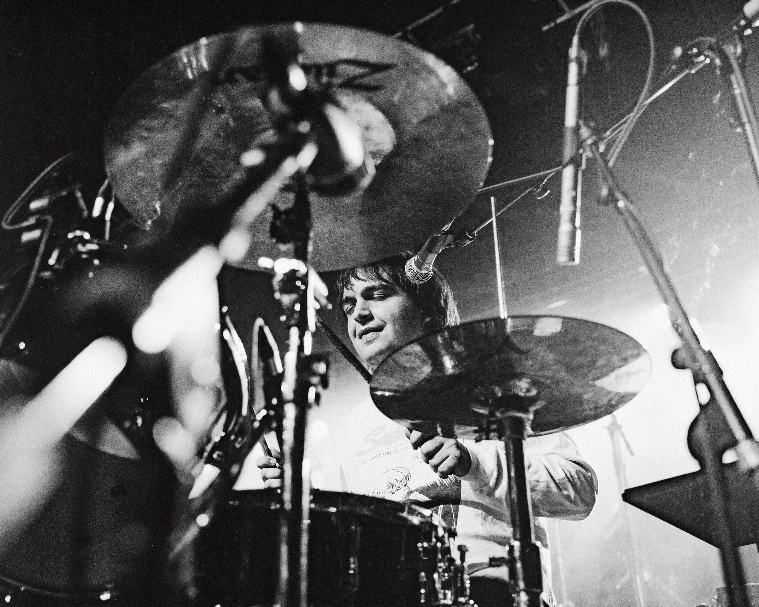 A black and white photo of a musician playing drums on stage, seen from underneath the cymbals with a close-up of their face and hand.