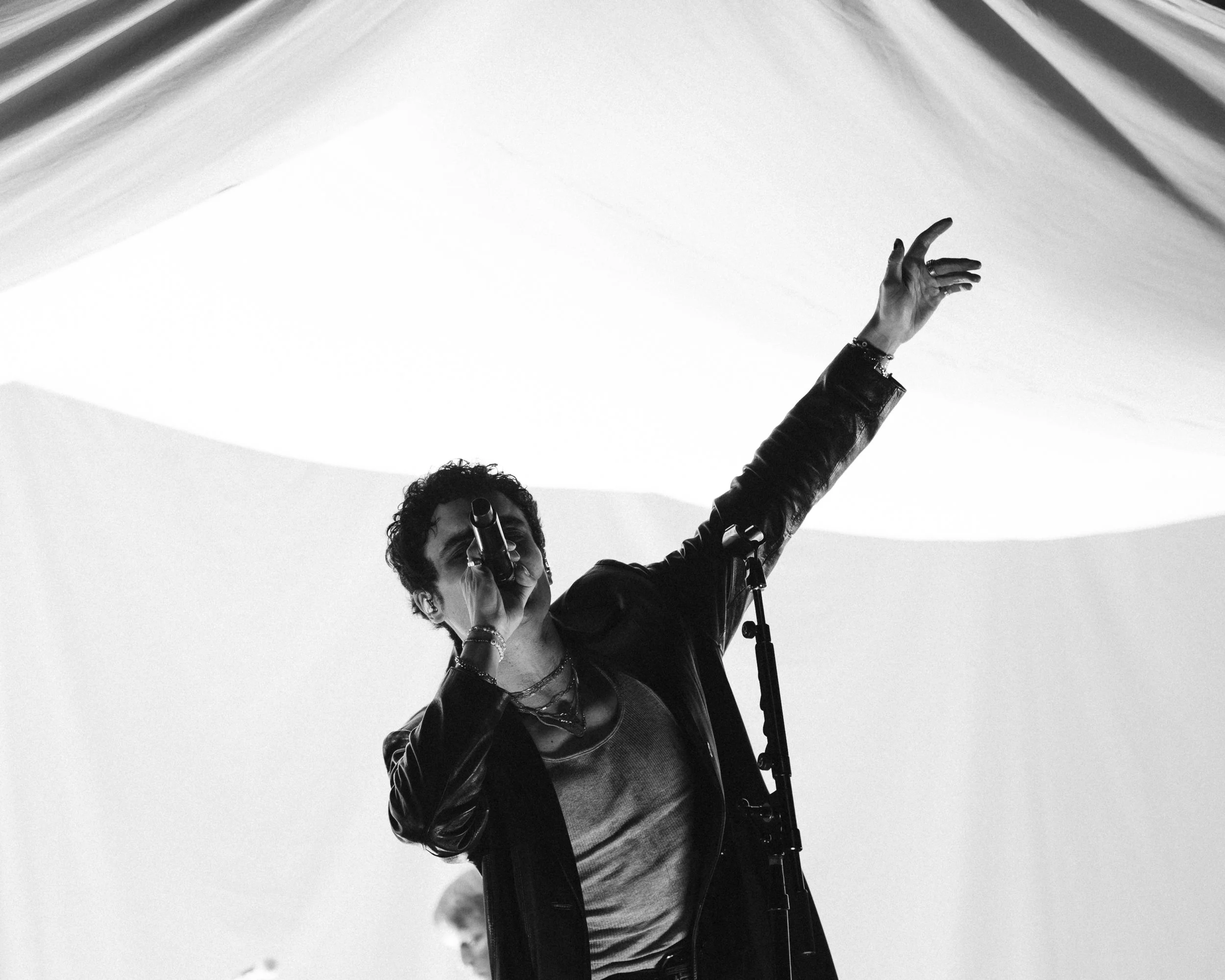 A black and white photo of a person singing into a microphone with their arm raised, standing in front of an abstract background.