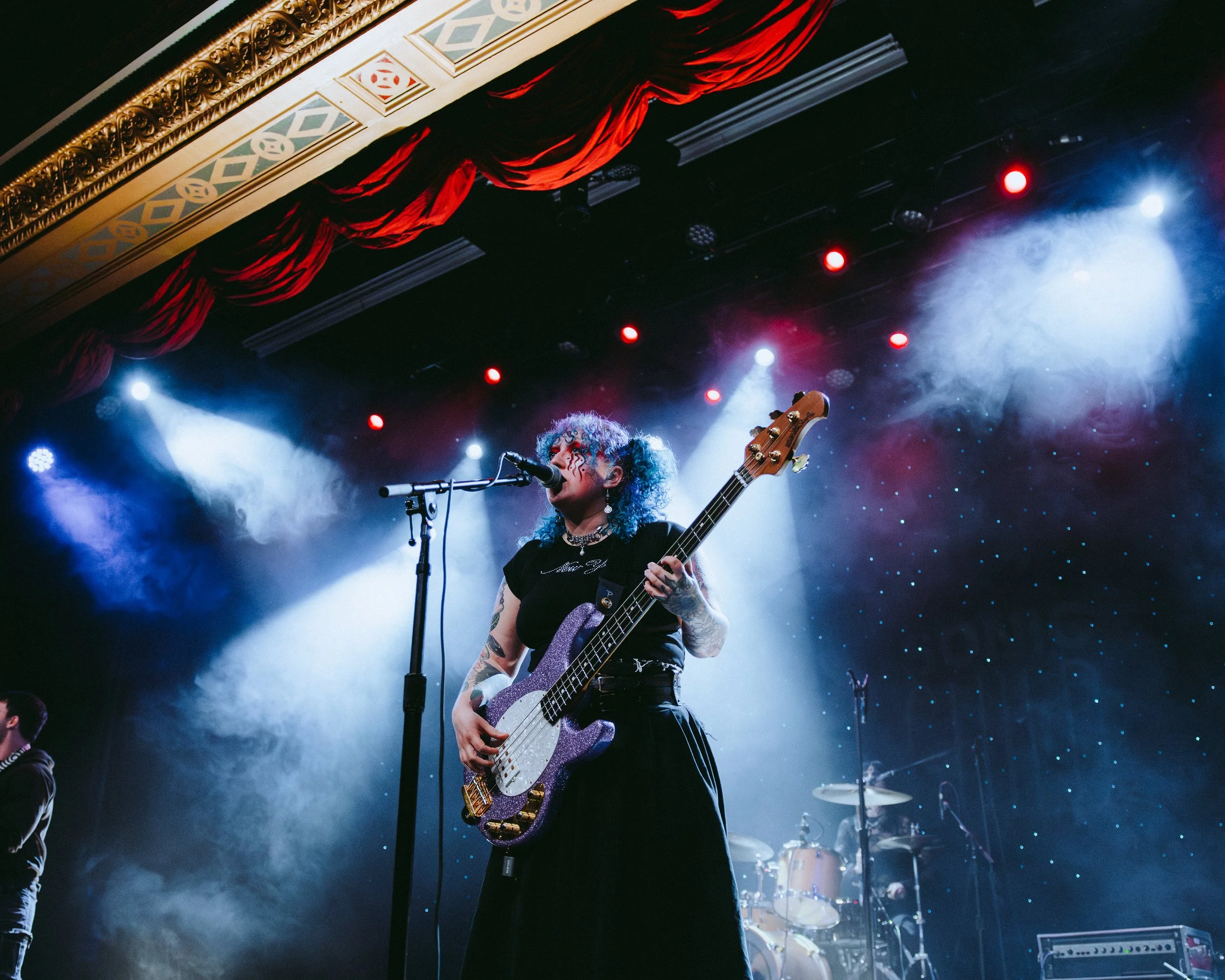 A woman with curly hair playing a purple bass guitar on stage with drums in the background and colorful stage lighting.