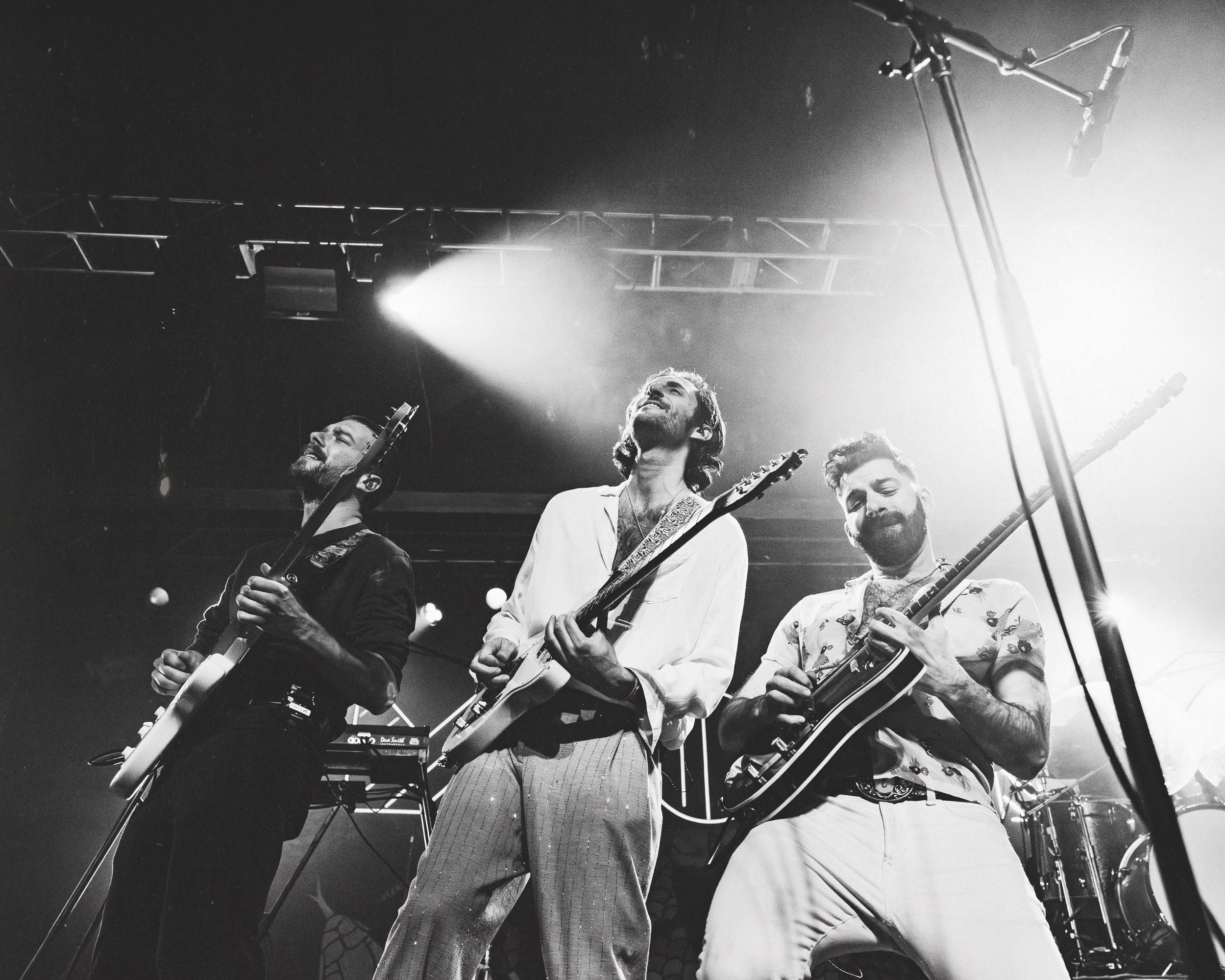 Three male musicians playing electric guitars on stage during a live concert, with stage lights and microphone setup visible in black and white.