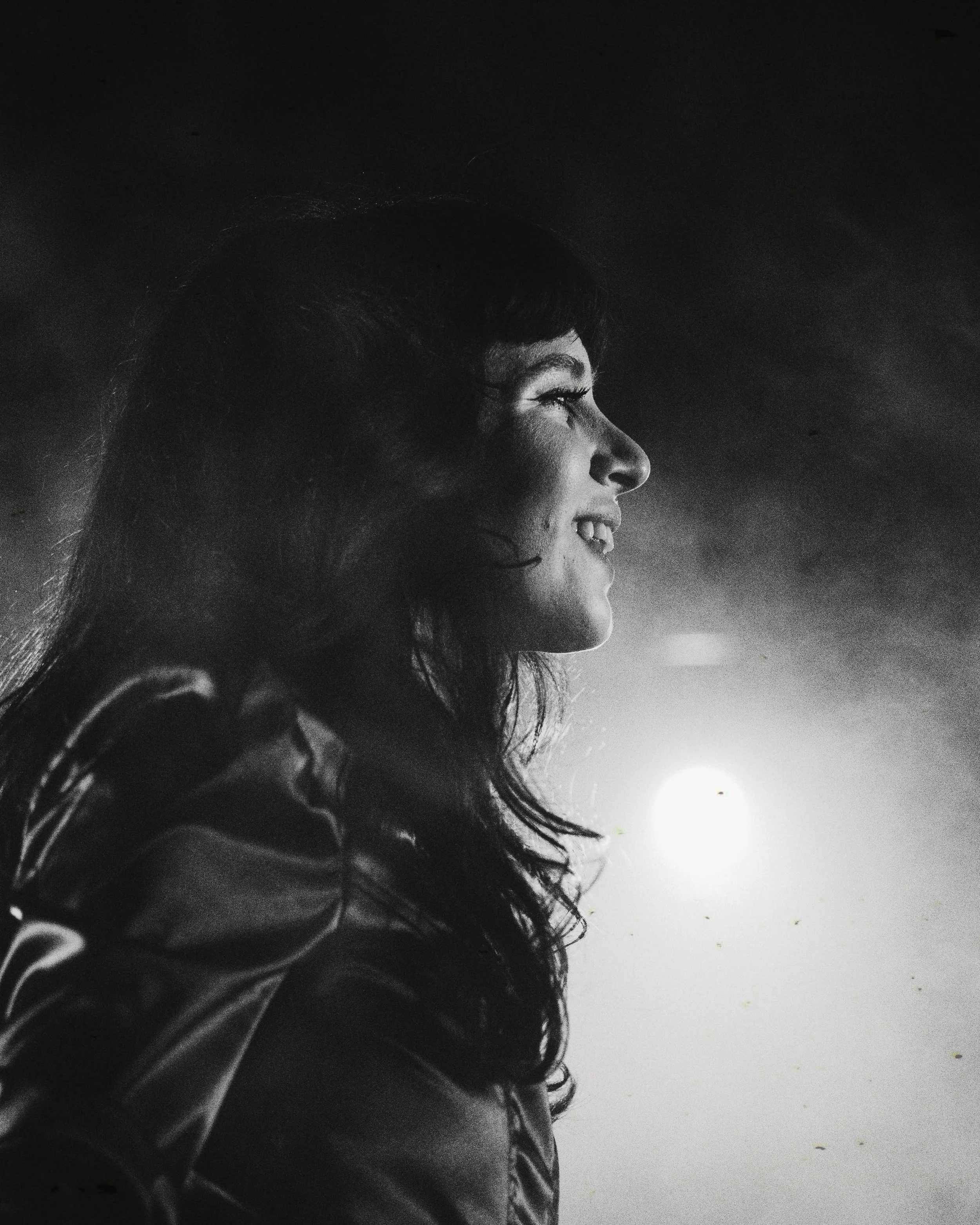 Side profile of a smiling woman with dark hair, illuminated by a bright light behind her.