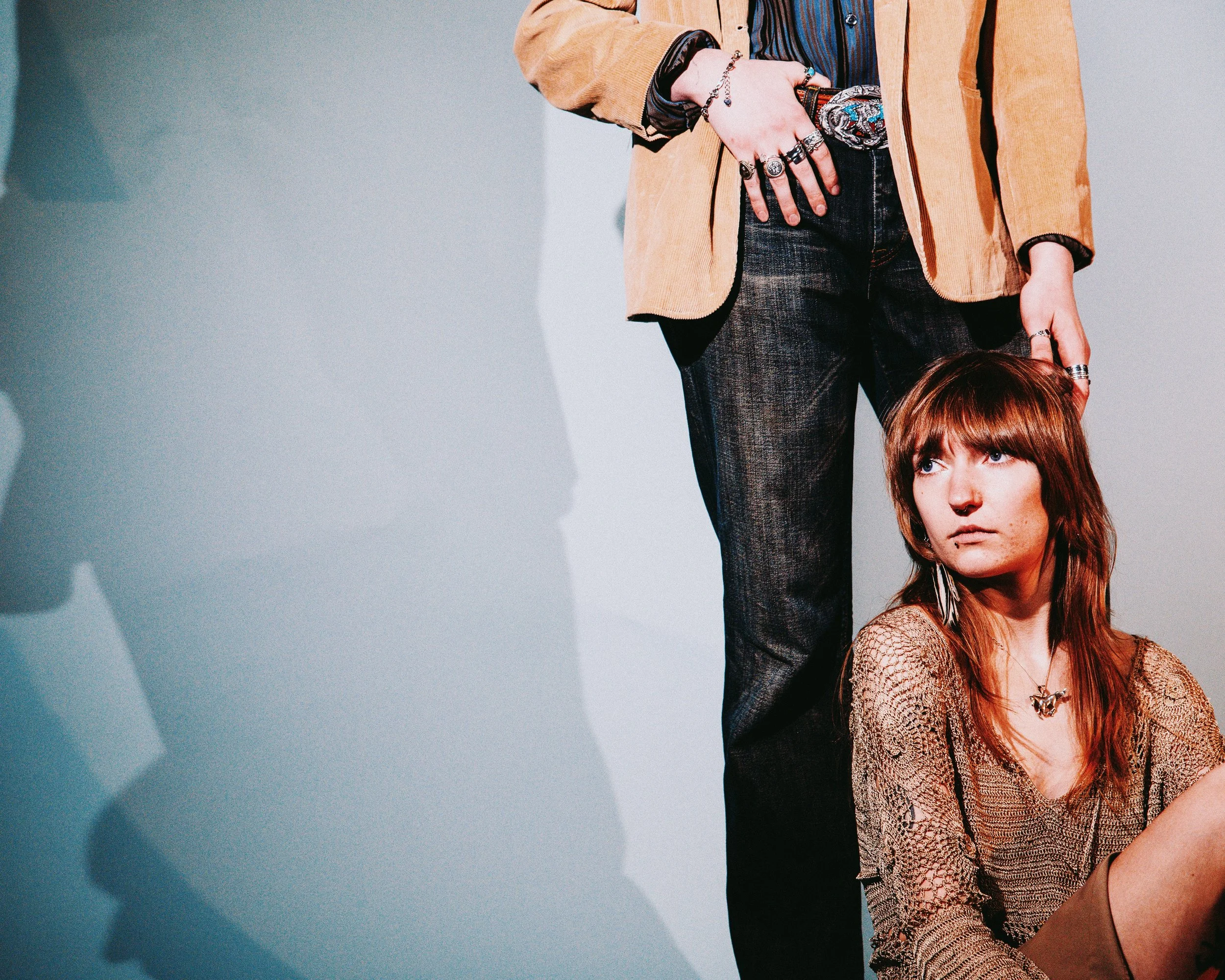 A woman with red hair sitting on the floor, looking serious, wearing a beige patterned top, with her head slightly tilted to the side. A person standing nearby is only partially visible, wearing a tan blazer and dark jeans. The person has multiple ri