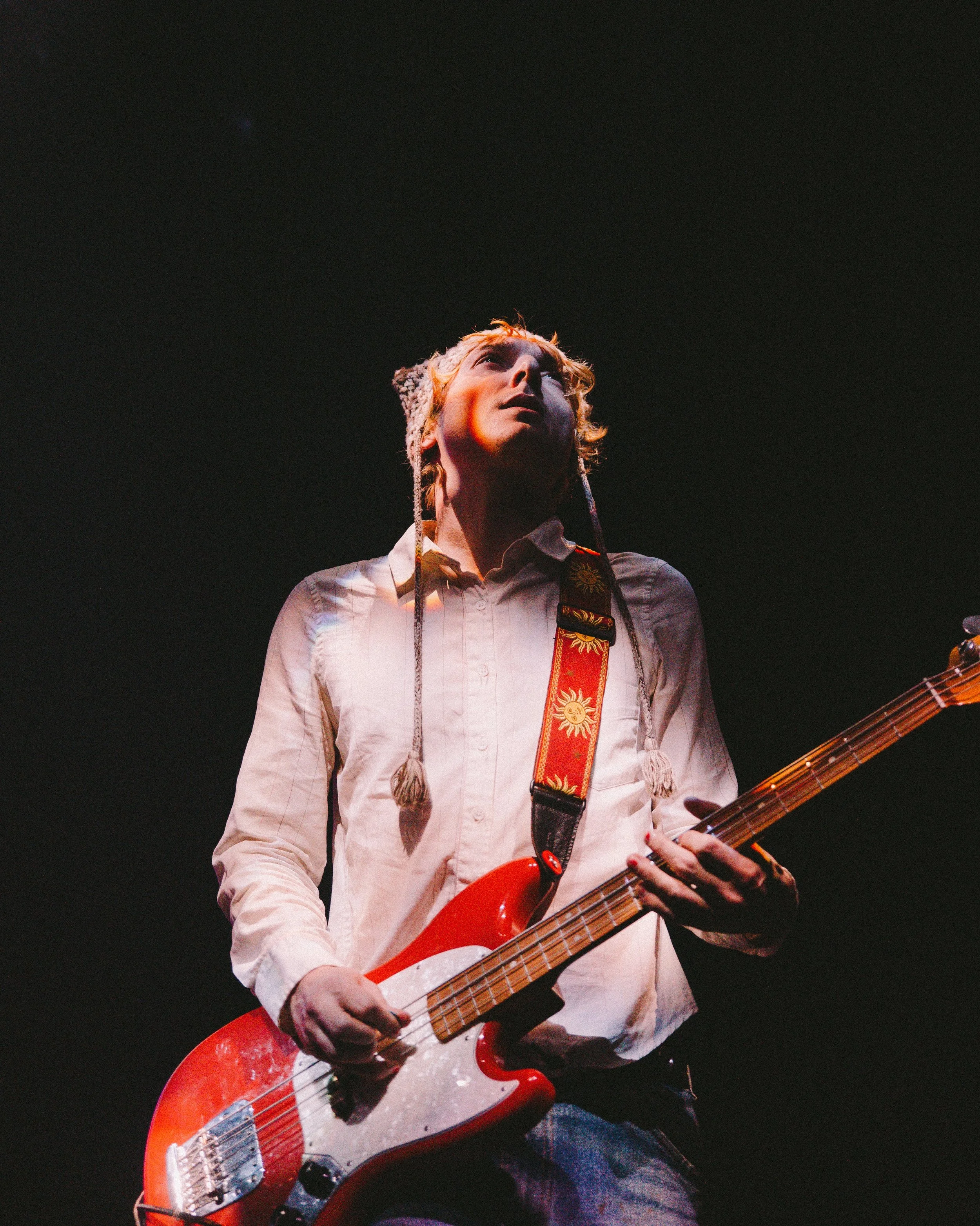 A person with a beige button-up shirt playing a red electric bass guitar on stage, with a dark background.