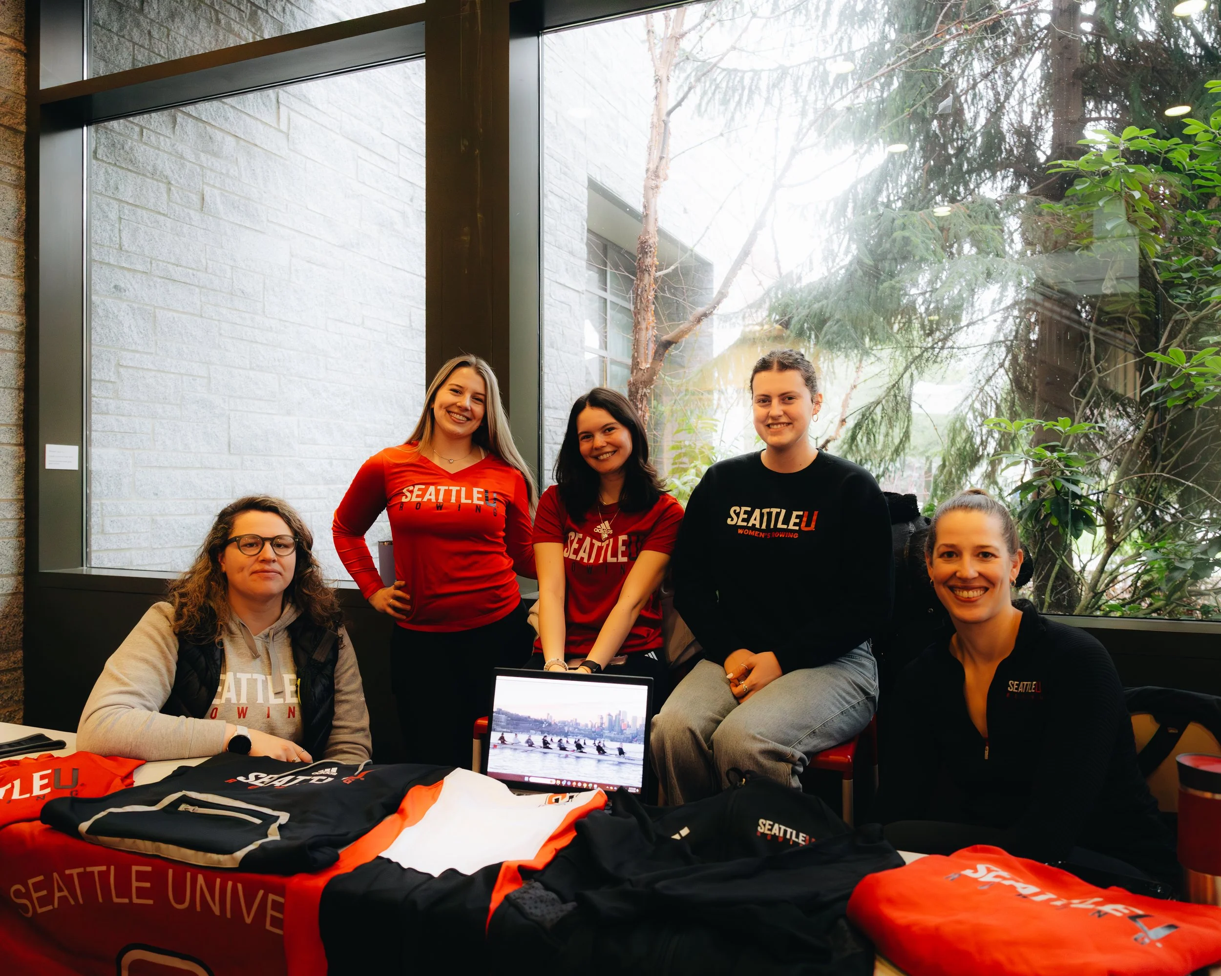Five women sitting and standing behind a table with Seattle University merchandise, including jackets and sweatshirts, in front of a large window with trees outside.