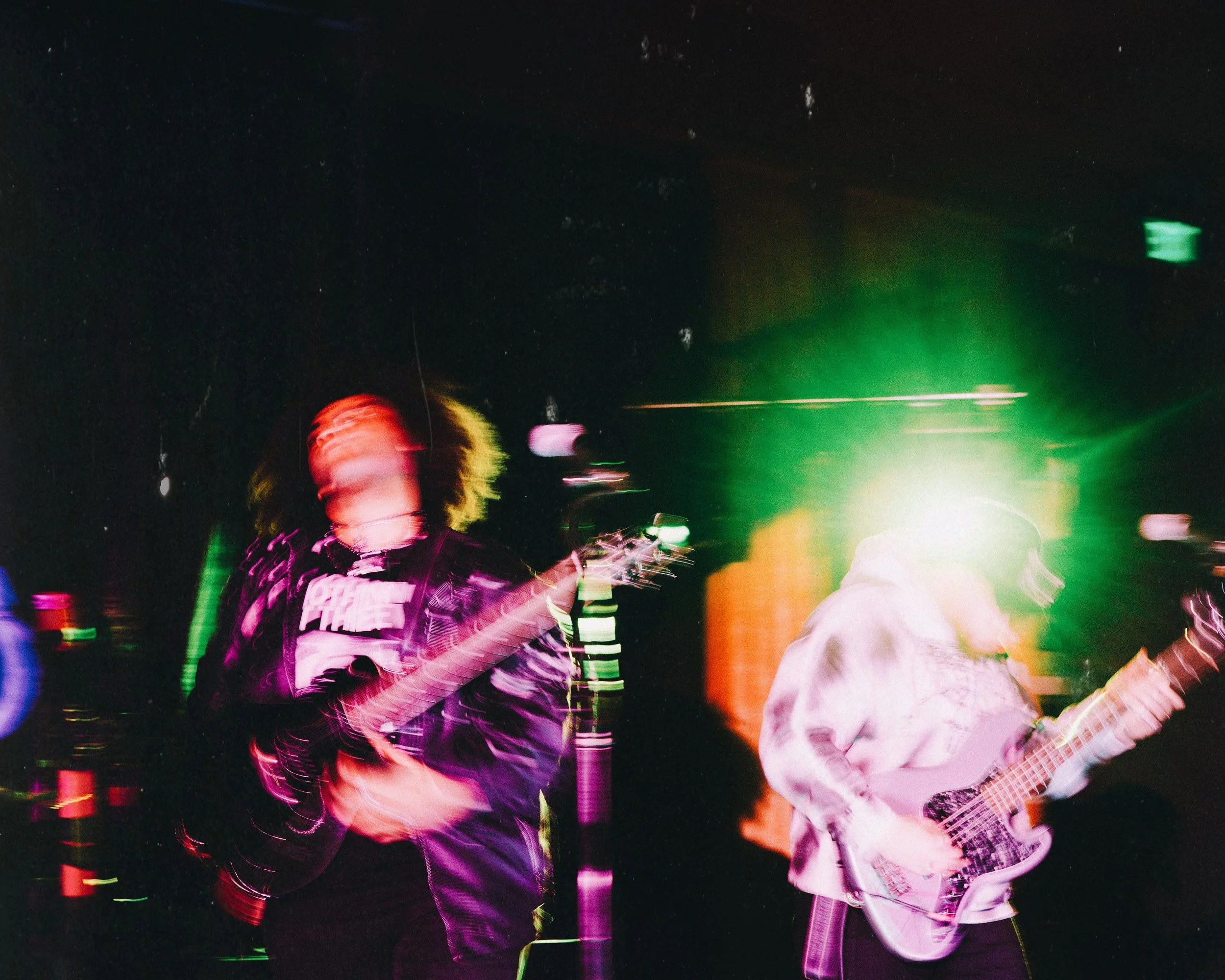 Two musicians playing electric guitars on stage with colorful light effects and motion blur.