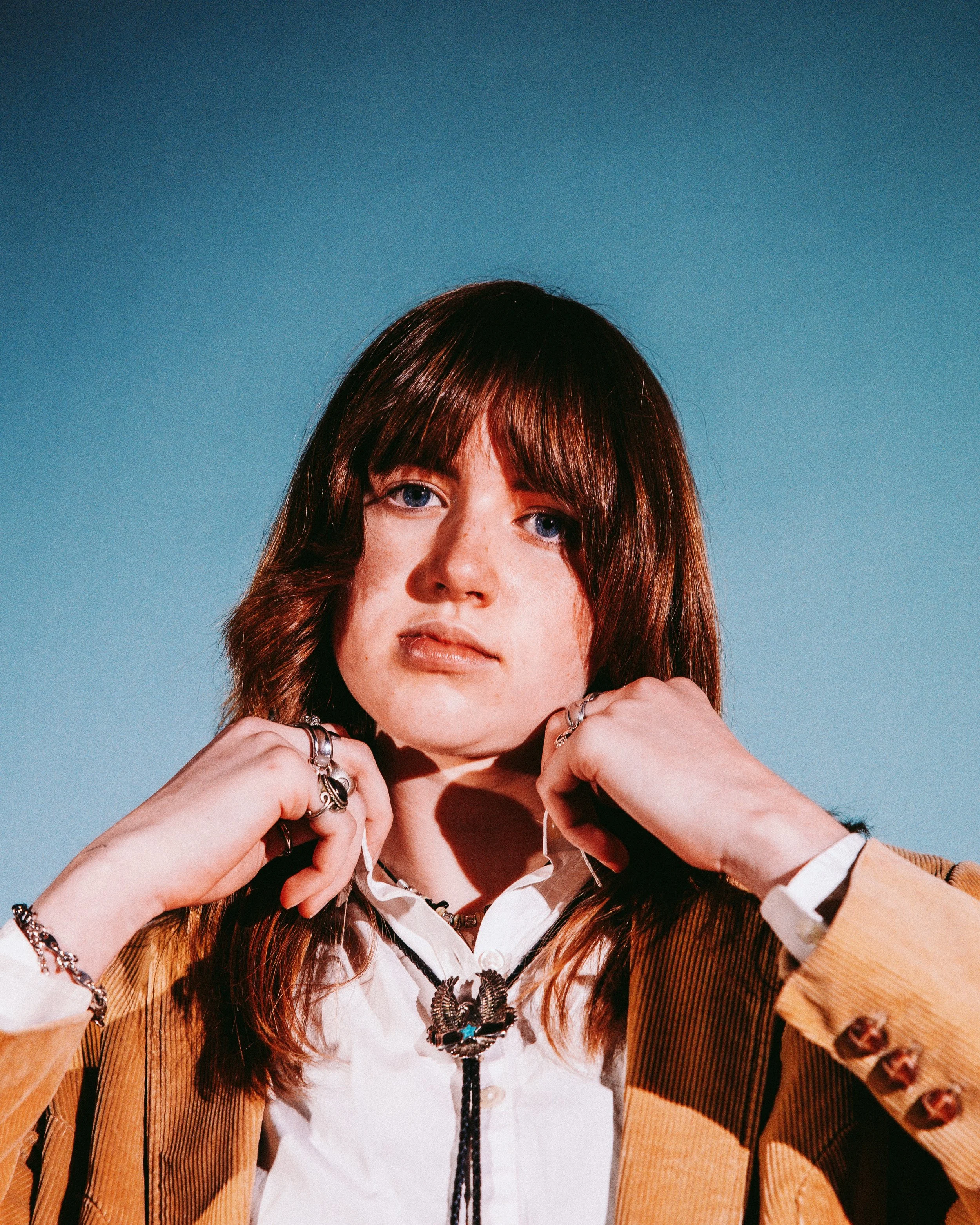 A young woman with red hair and blue eyes, wearing multiple rings and jewelry, posing against a blue background.