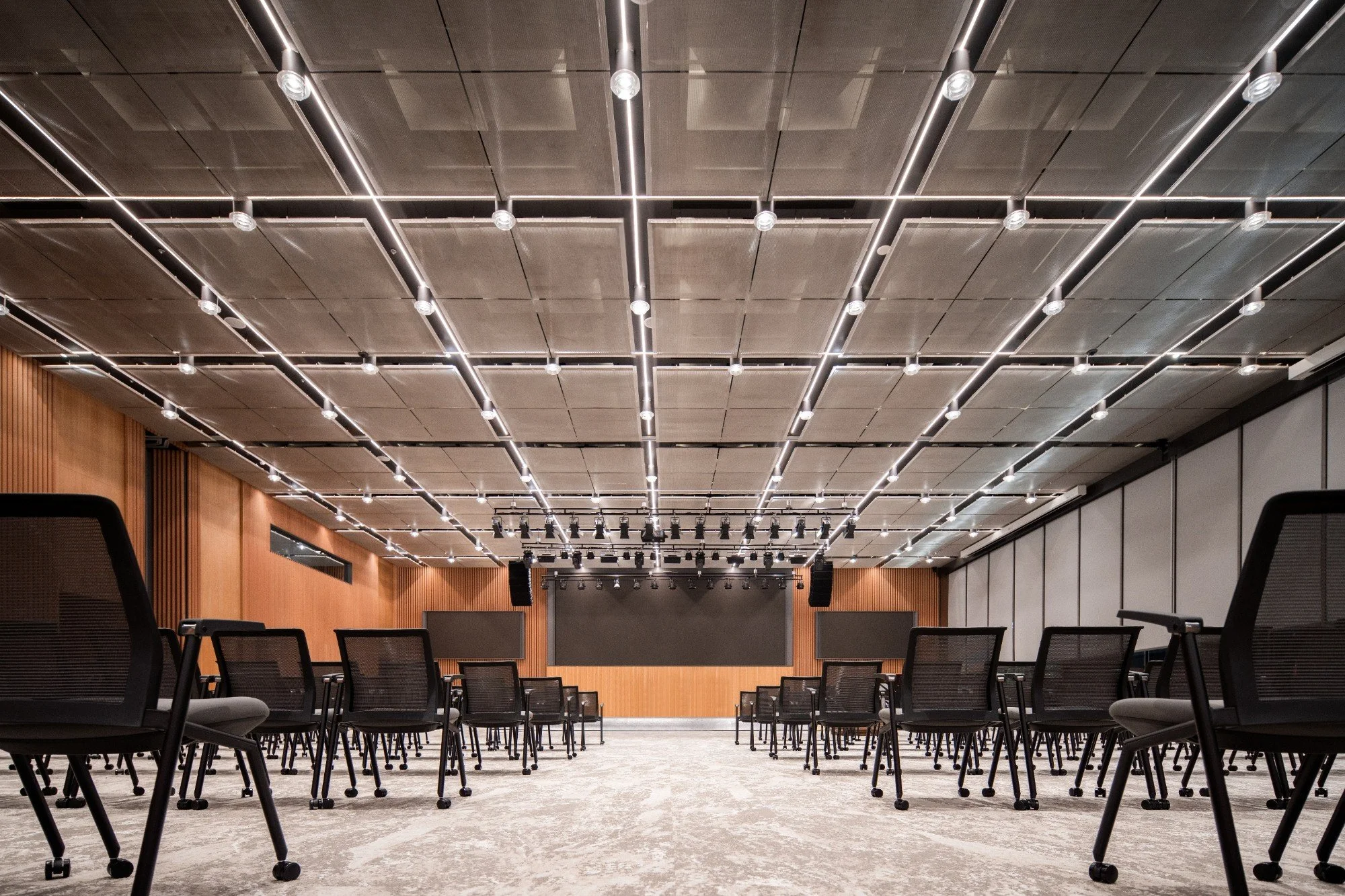 Large conference room with chairs arranged facing a stage, featuring a large screen and professional lighting overhead.