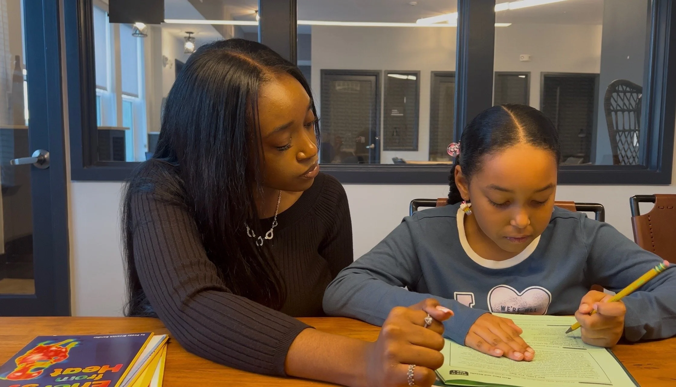 An adult woman and a young girl sitting at a table, looking at a booklet or worksheet together. The woman is wearing a black top and has long black hair, while the girl has braided hair with hair accessories and is holding a yellow pencil.