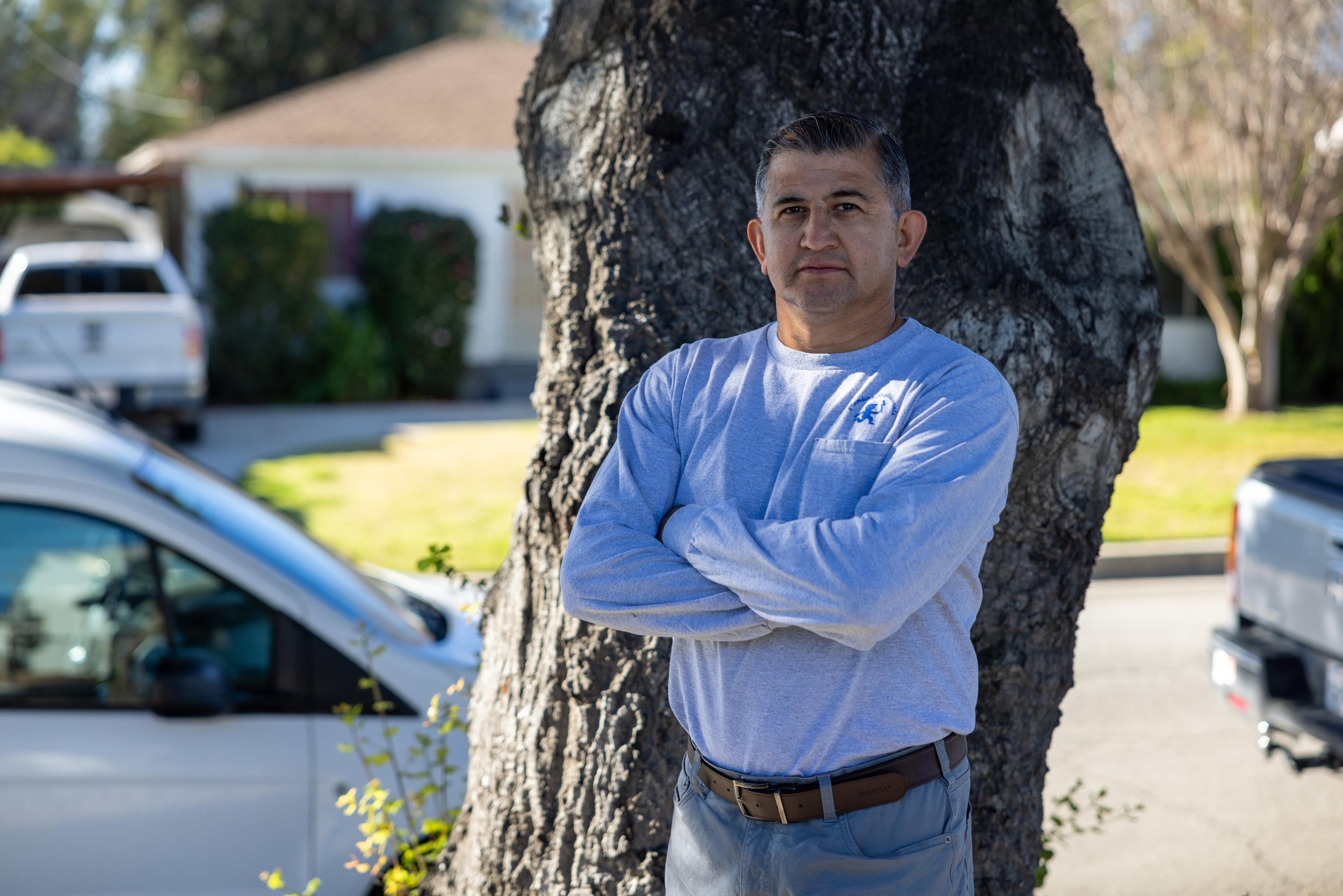 A man with gray hair wearing a gray long sleeve shirt and dark jeans, standing with crossed arms and leaning against a tree in a suburban neighborhood. There are parked cars and houses in the background.