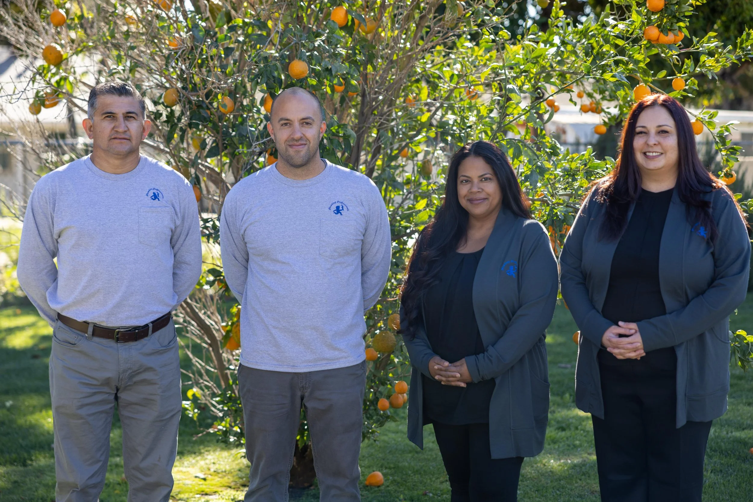 Four people standing outdoors in front of an orange tree with ripe oranges. They are smiling and wearing casual clothing.