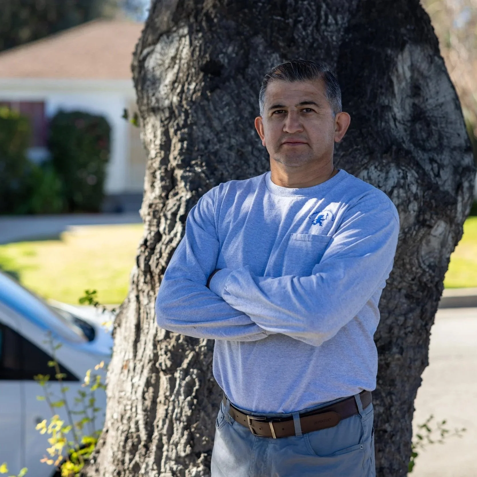 A middle-aged man with dark, slicked-back hair and light skin stands outdoors near a large tree, with arms crossed, looking into the camera. He is wearing a light gray long-sleeve shirt with a small logo on the chest and a brown belt. In the blurred background, there are houses, greenery, and a car, suggesting a suburban neighborhood.