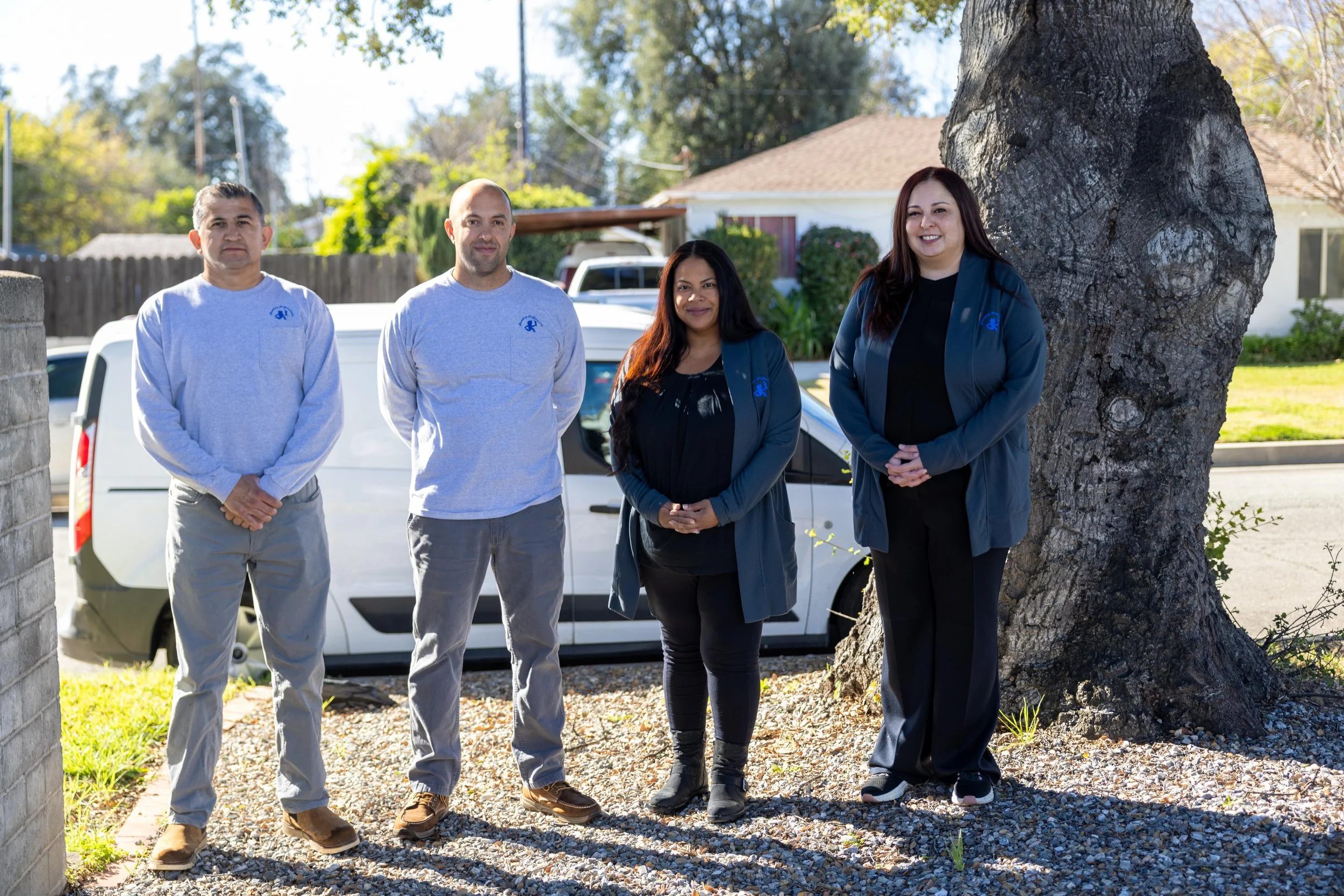 Group of four diverse people standing outdoors near a tree, smiling, with a white vehicle and residential neighborhood in the background.