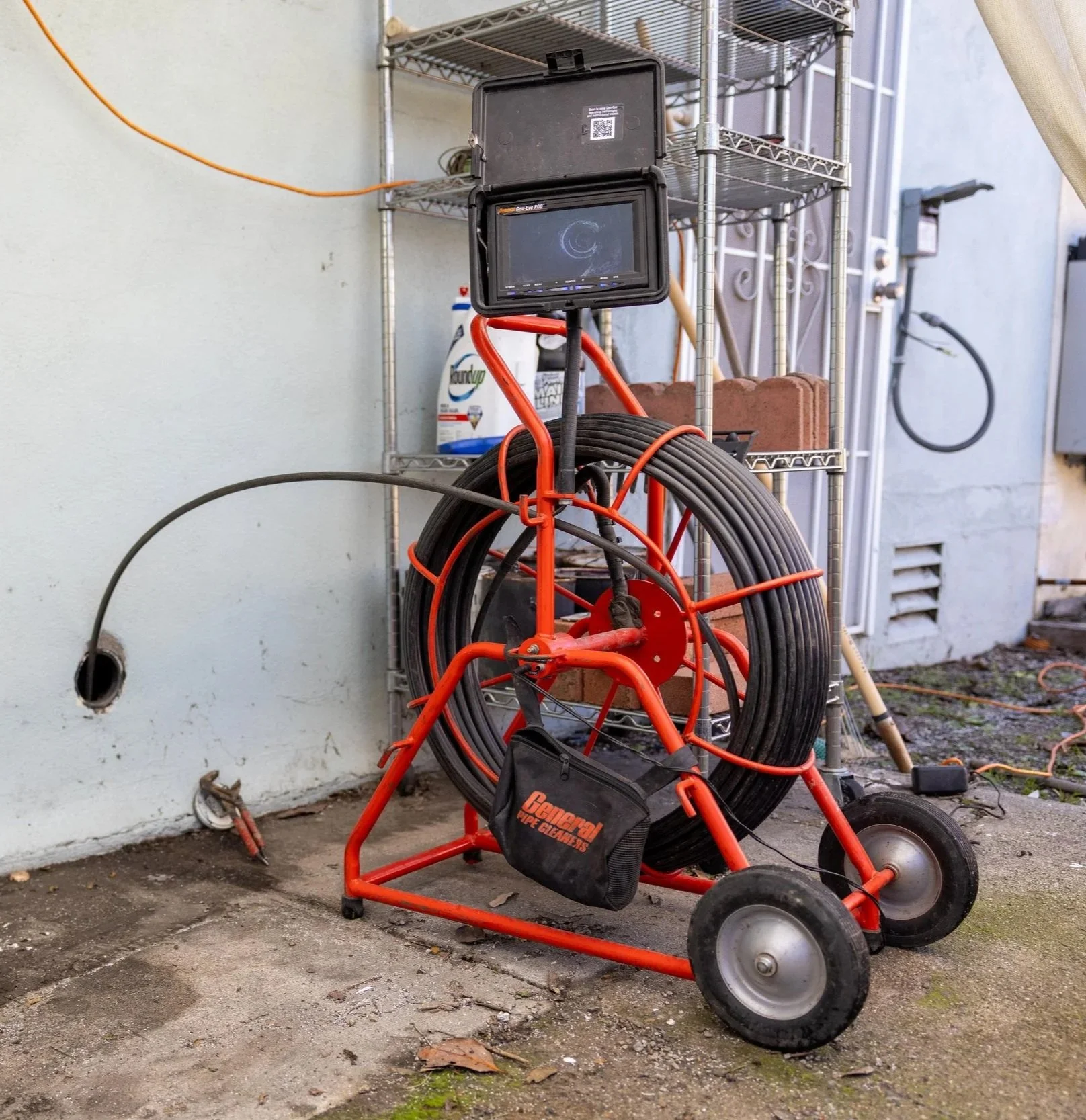 Orange pipe inspection camera cart with a monitor, a large coil of hose, and a black bag labeled 'General Pipe Cleaners' outside on a concrete surface by a white wall and metal shelving.