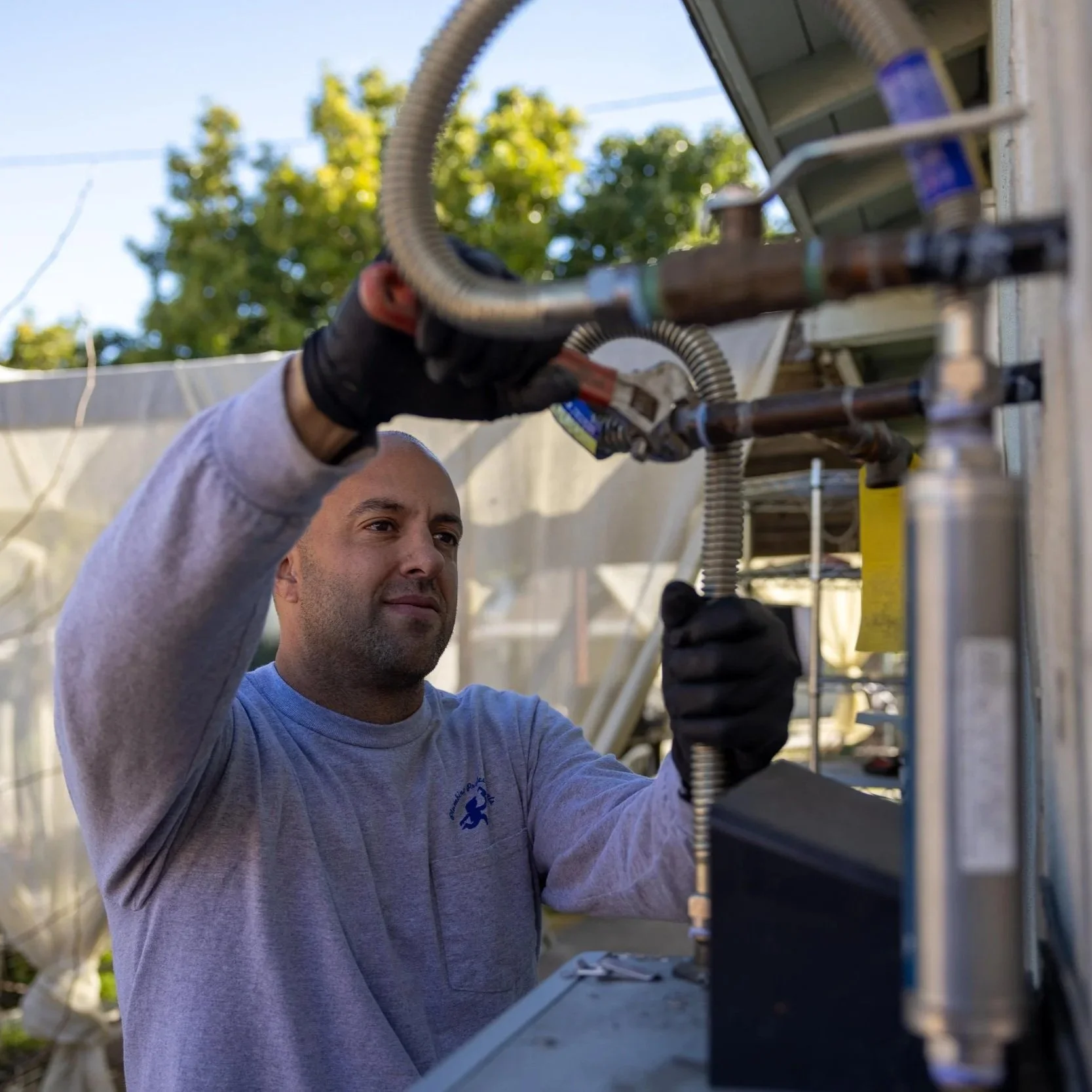 A man working on an outdoor electrical or communication box, wearing gloves and using tools, with trees and a clear sky in the background.