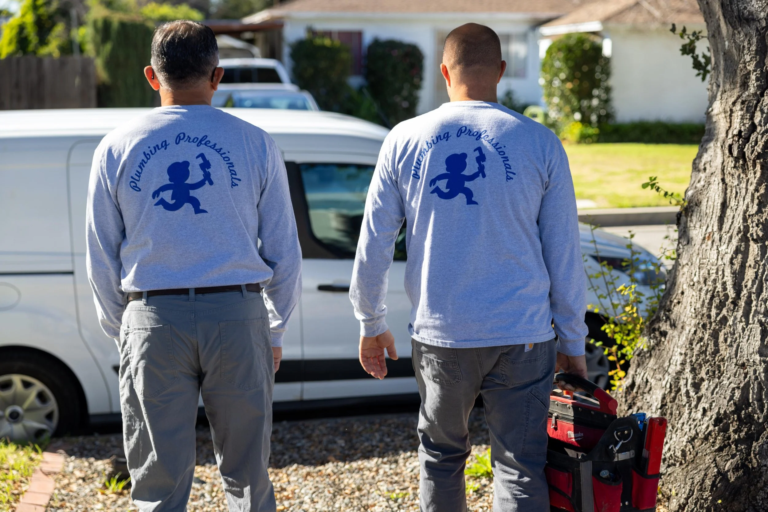 Two male plumbers in gray shirts with a cartoon plumber logo, standing outdoors near a white van and tree, preparing tools for work.