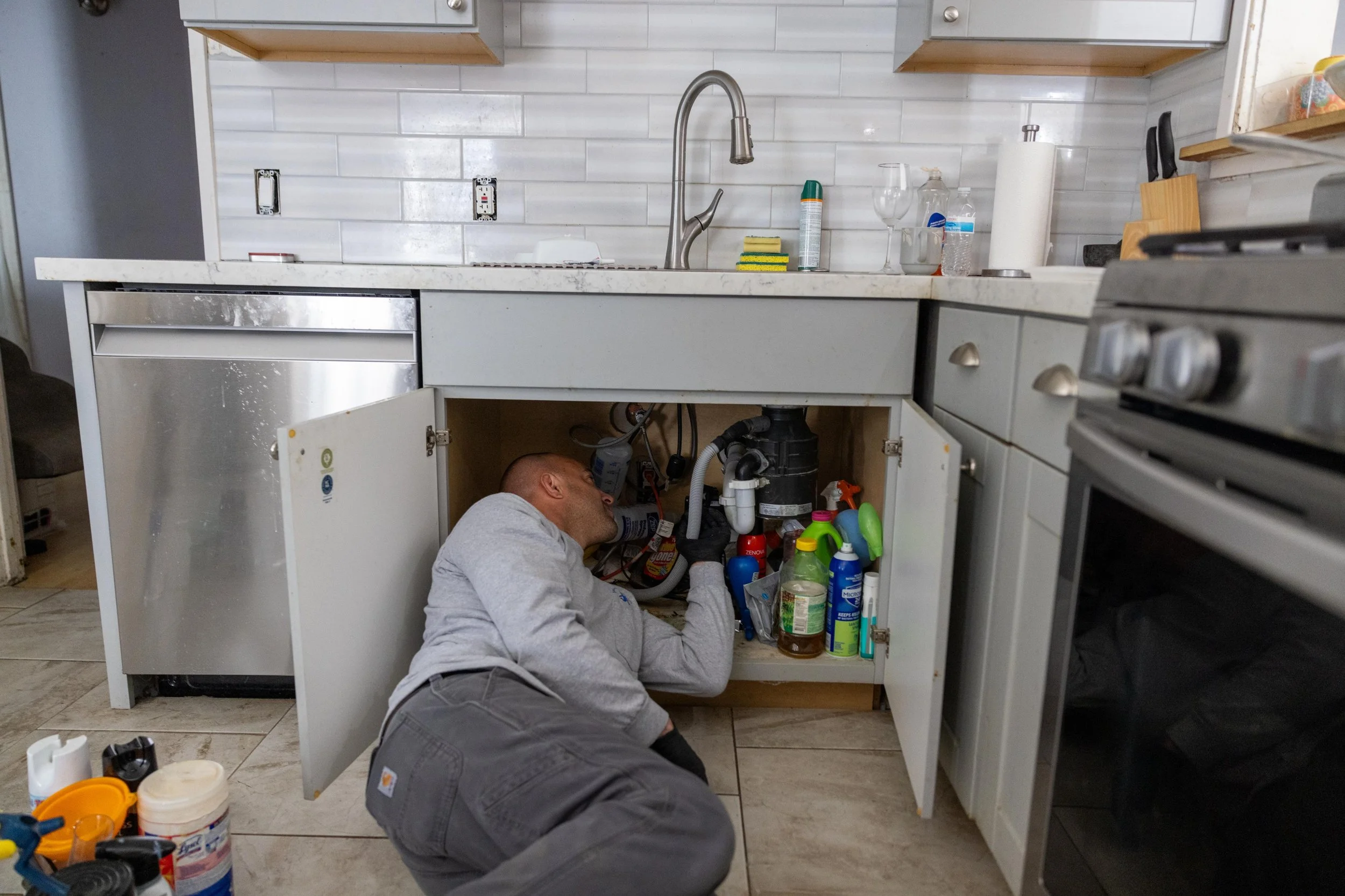 A man working underneath a kitchen sink cabinet, repairing or inspecting plumbing. He is lying on the floor with tools and supplies around him, in a kitchen with a white tiled backsplash, a stainless steel faucet, and various cleaning products and paper towels on the countertop.