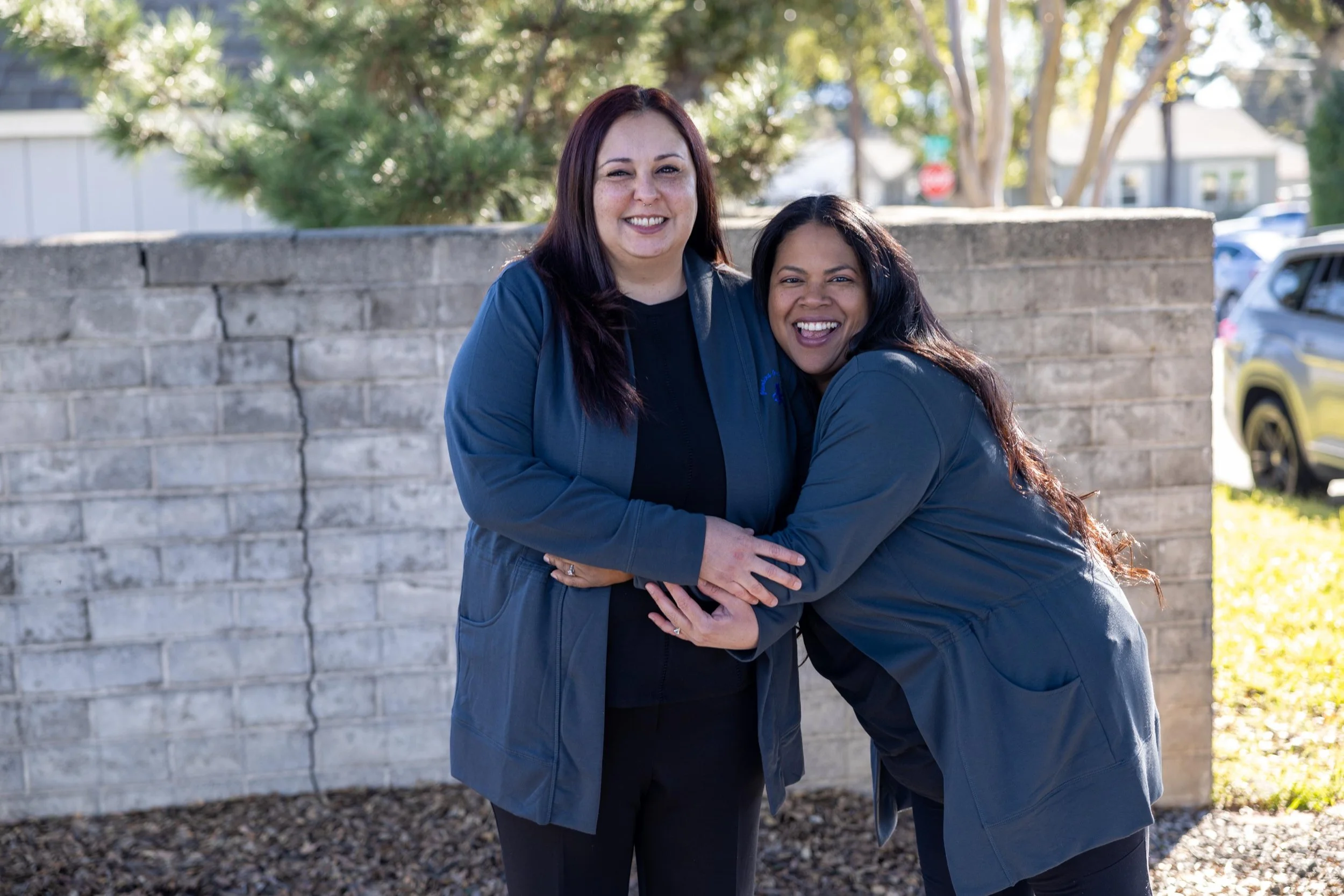 Two women smiling and hugging outside near a stone wall, with trees and cars in the background.
