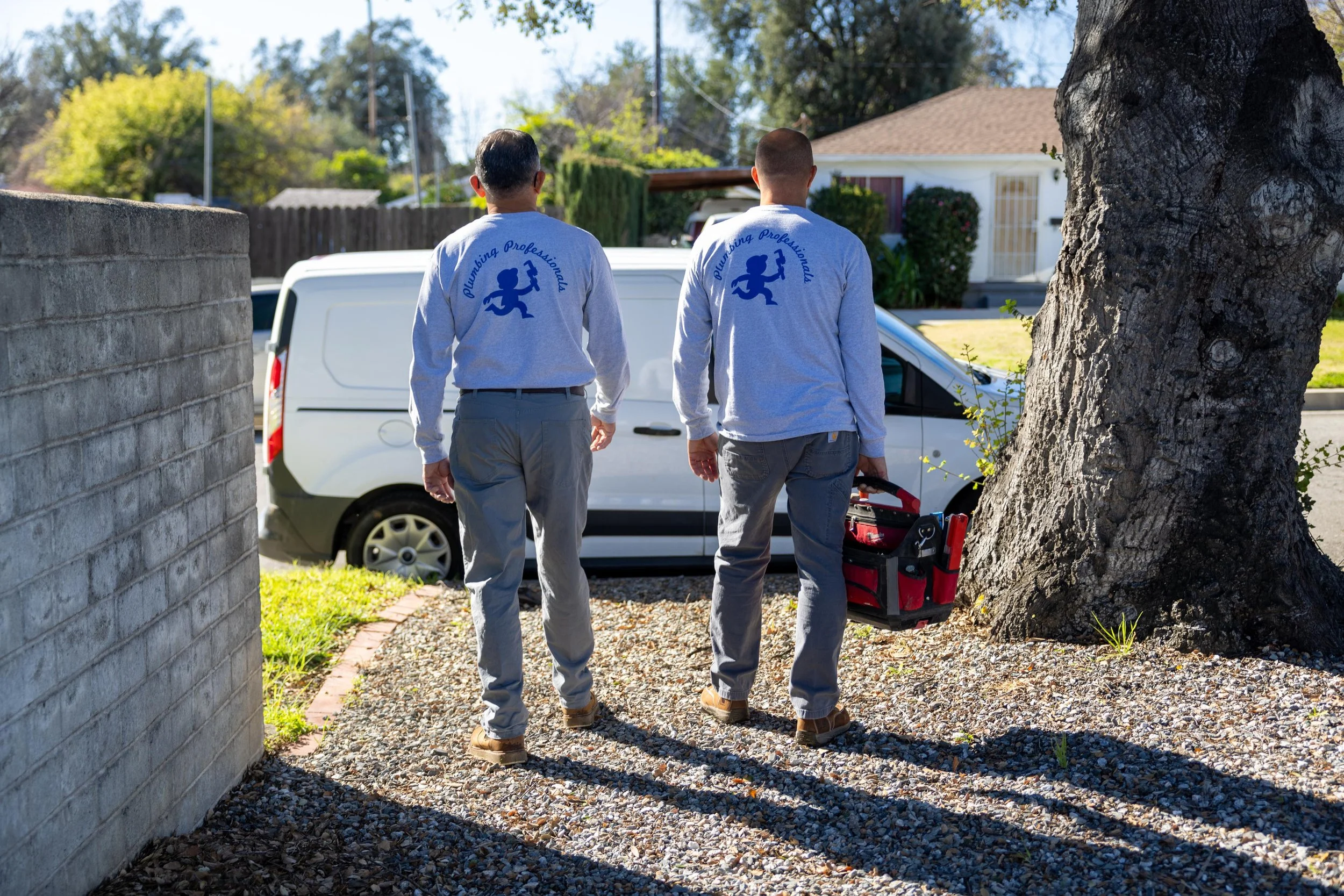 Two plumbing professionals wearing matching gray shirts with a bear logo and the words "Plumbing Professional" on the back, walking on a gravel path in a residential neighborhood, carrying a toolbox in one hand.