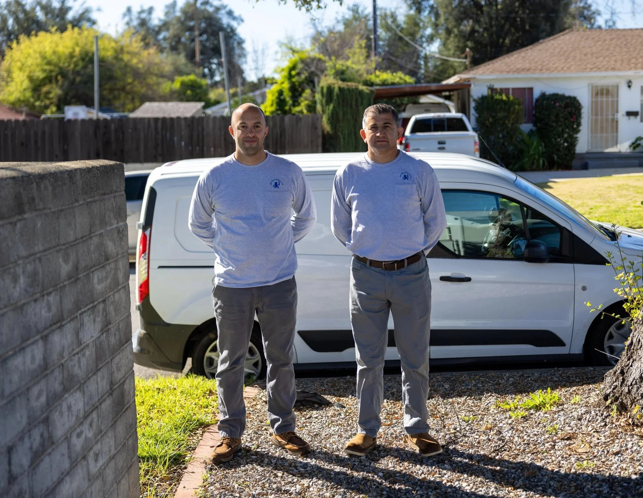 Two men stand outdoors in front of a parked white van, wearing matching light gray shirts with a small logo. Background includes a house, a wooden fence, and trees under a clear sky.
