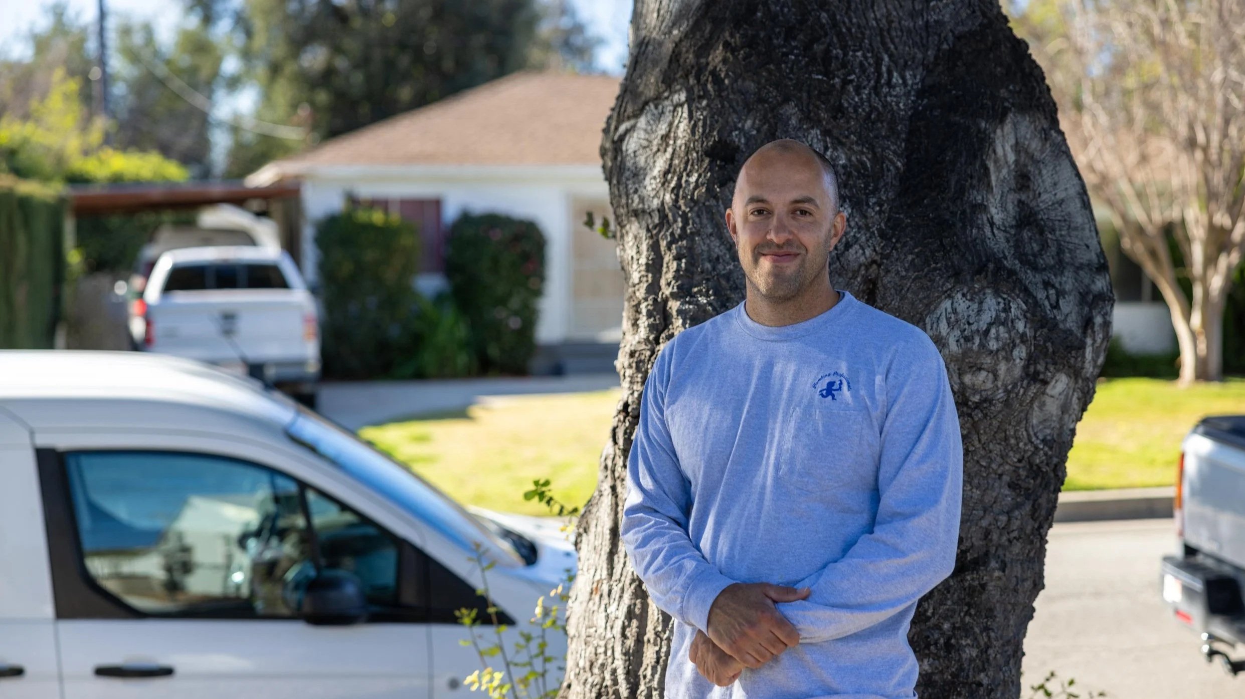 A man with a bald head and slight beard standing outdoors next to a large tree, wearing a light blue long-sleeve shirt, with cars and a house in the background.
