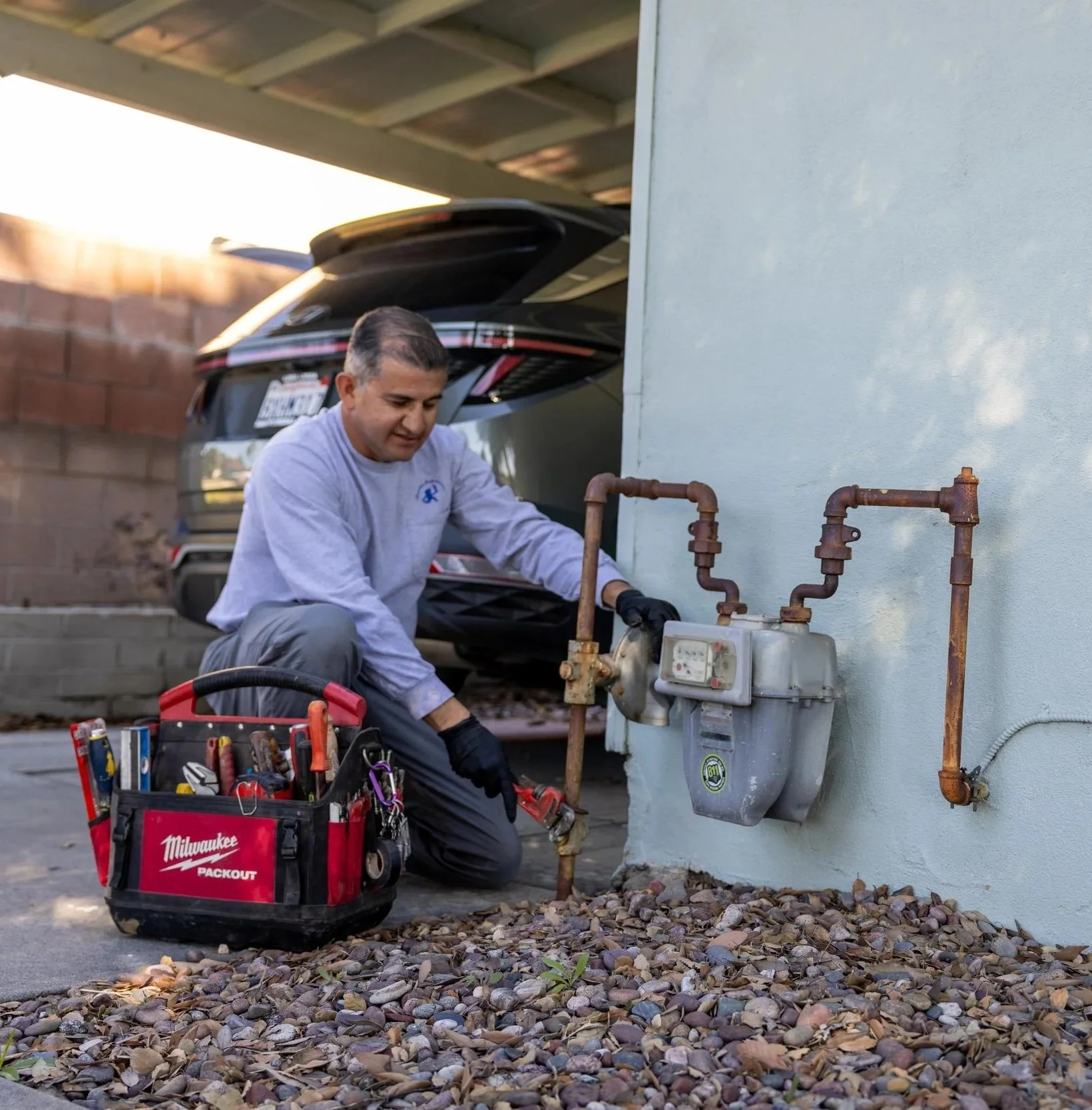 A handyman working on plumbing outside near a house with a black car parked in the background. He is kneeling on rocks, using tools from a red Milwaukee tool bag, and wearing a gray shirt and black gloves.